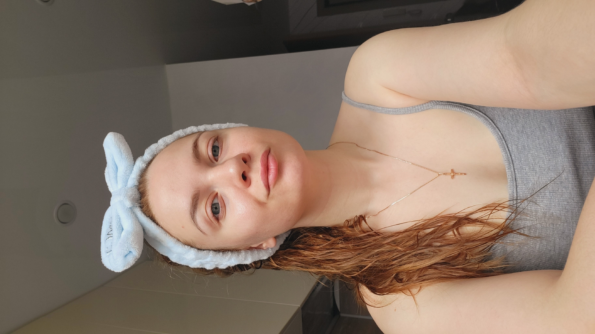 A young woman is lying down, facing upward. She is wearing a gray tank top and a light blue headband with bunny ears. She has reddish-brown, wavy hair that appears wet. A gold necklace with a cross pendant is visible on her chest. The background is a wall and ceiling, with what appears to be a light fixture visible. No information about Chaikovsky, Russia can be gleaned from the image.