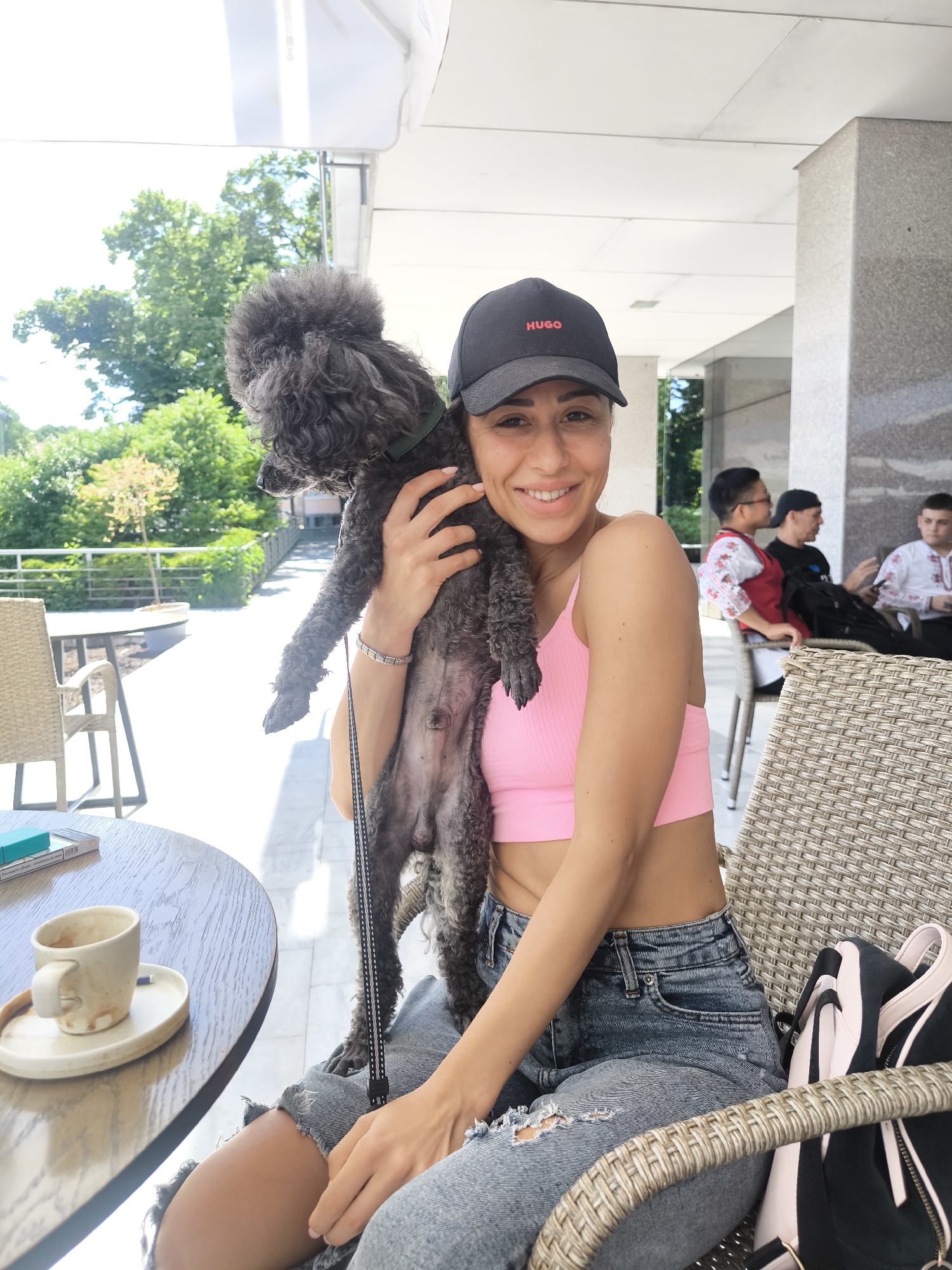 The image shows a woman seated outdoors at a café in Stara Zagora, Bulgaria. She is wearing a pink cropped top, ripped denim shorts, and a black "HUGO" baseball cap. She is holding a black poodle close to her body. On the table next to her is a dirty, half-finished cup of coffee. In the background, under a covered outdoor seating area, there are several people seated at a table, including two young men wearing traditional embroidered shirts. The area is surrounded by greenery and buildings. A black and pink backpack hangs behind the woman’s chair.