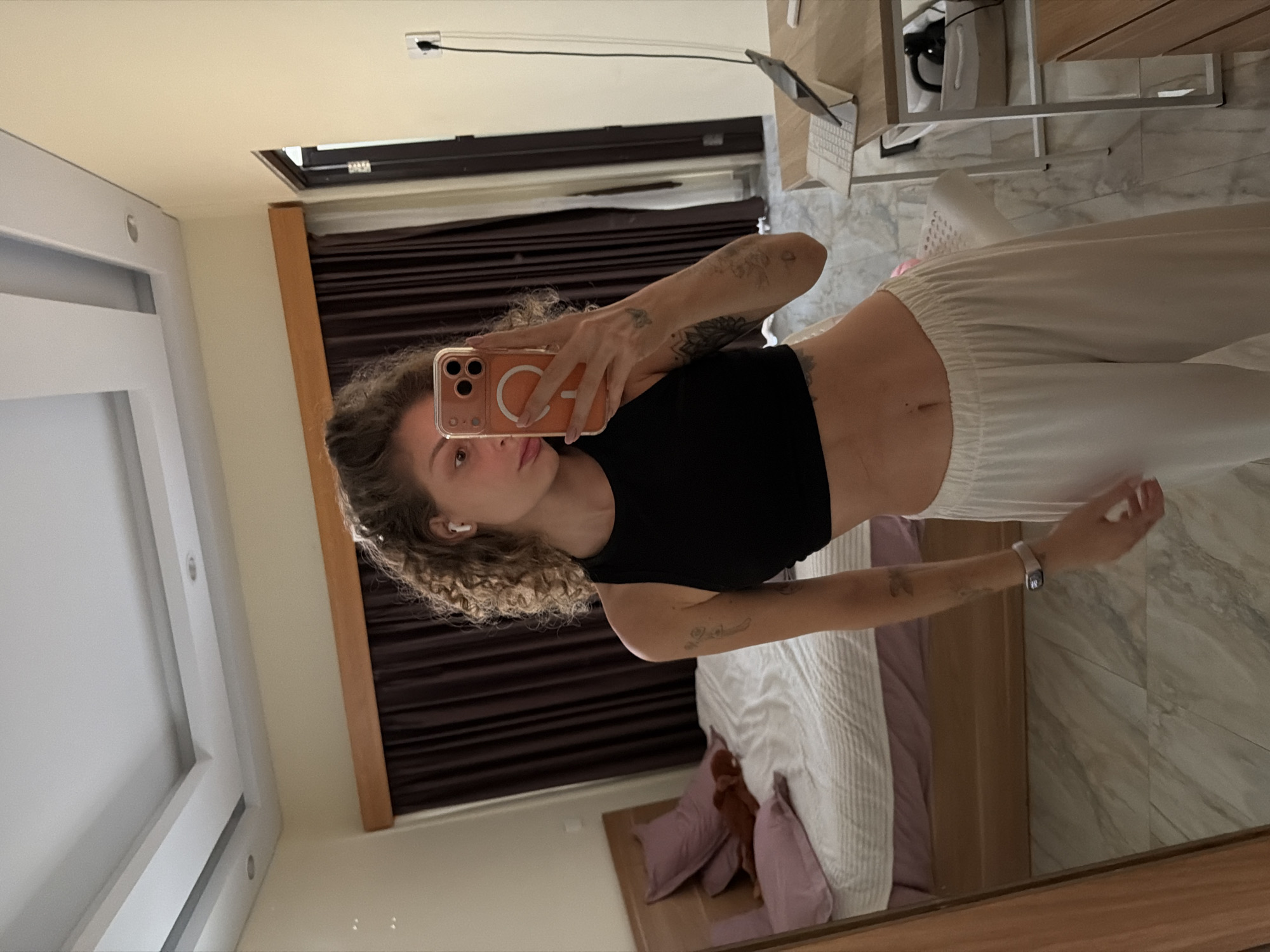 A mirror selfie captures a young woman standing in what appears to be a bedroom or studio apartment, located in North Kuta, Indonesia. The image is oriented sideways, but depicts an upright view of the scene.
The woman is the central subject, holding a light orange iPhone with a MagSafe ring on the back, covering part of her face as she takes the photo. She has curly, light brown hair and is wearing a black crop top that reveals her midriff, paired with loose-fitting, light-colored pants. Tattoos are visible on both of her arms, and she wears white wireless earbuds. A watch adorns her left wrist. Her expression is neutral, as she looks directly into the phone camera.
The background, reflected in the mirror, shows an indoor setting. The floor is covered in light-colored marble or tiles with grey veining. A bed with a light-striped duvet and two purple pillows is visible. Dark brown curtains are drawn over a window, suggesting privacy or light control. To the right, a wooden desk or shelf holds a silver laptop with a visible keyboard, and possibly headphones or a bag. The walls are painted a light cream or beige color, and a light fixture or vent is seen on the ceiling. The overall lighting is bright and even, indicating daytime.