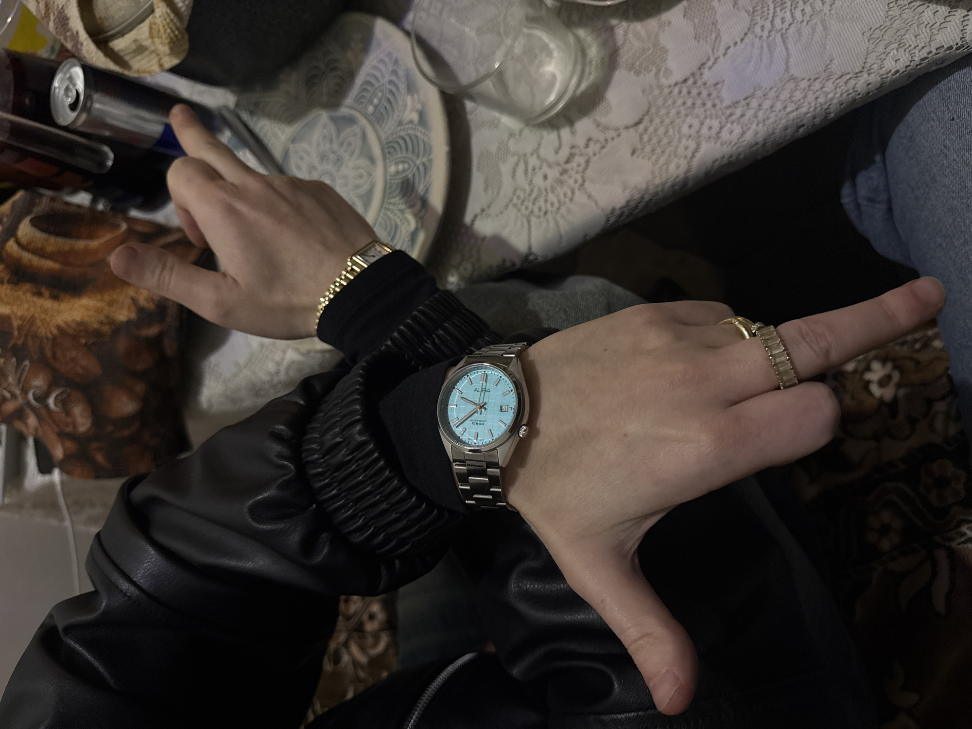 The image depicts a person sitting at a table with a floral patterned tablecloth. The person is wearing a black leather jacket and jeans. They are wearing two watches, one with a light blue face and a silver band on the right wrist, and a gold watch on the left wrist, along with a gold ring on the right hand. The hands are positioned in a gesture, with index fingers extended. Objects on the table include a plate, a glass, and cans. The location is likely indoors, and there is no information to specifically pinpoint it in Yerevan, Armenia.
