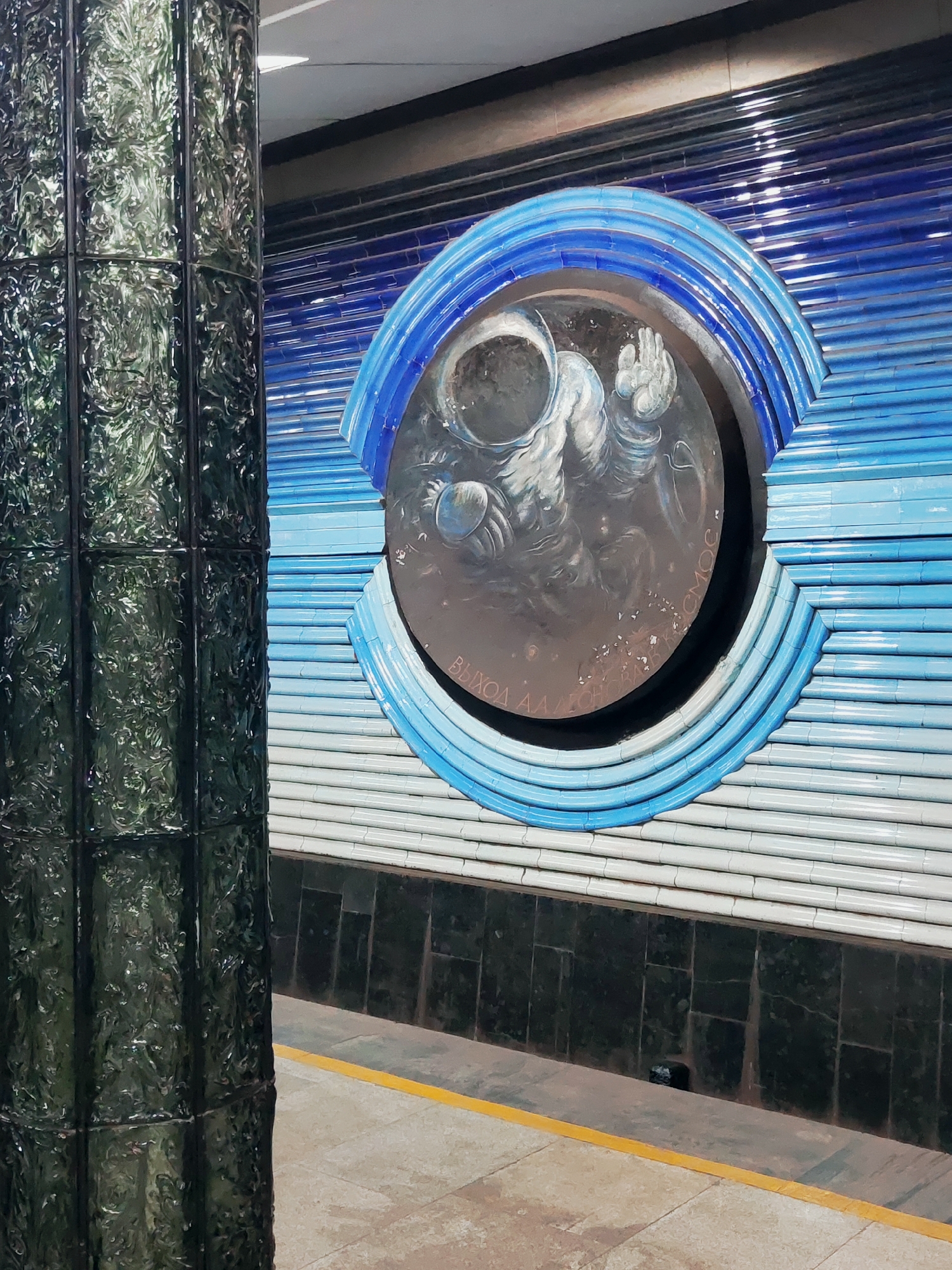 The image shows a mural of an astronaut within a circular frame on a tiled wall, likely in a subway station. The mural is set against a backdrop of blue and white horizontal tiles. To the left of the mural, a dark, textured pillar stands prominently. The floor in front of the wall is paved, with a yellow line marking the edge of a platform. Text is visible within the circular frame of the mural, appearing to be in Cyrillic script. This artwork is located at the Kosmonavtlar metro station in Tashkent, Uzbekistan.