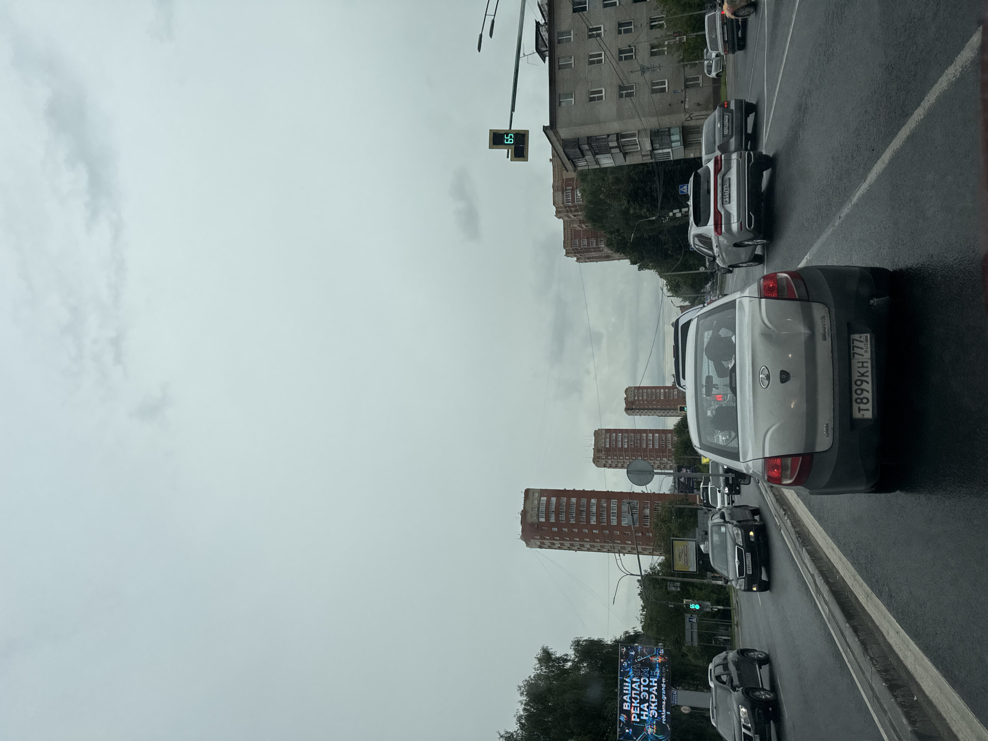 The image shows a view from inside a vehicle traveling on a wet asphalt road in Shchyolkovo, Russia. The sky is overcast and gray. In the foreground, the rear of a light-colored car is visible. The road has multiple lanes, and several other vehicles are present, including a dark SUV on the left and a silver SUV further ahead. Tall, multi-story brick buildings line the right side of the road. A traffic light displaying a green "65" is visible on a pole over the road. On the left side of the road, a large billboard with Russian text and graphics is displayed, accompanied by a dense line of trees. The overall scene depicts a typical urban street environment under cloudy weather conditions.