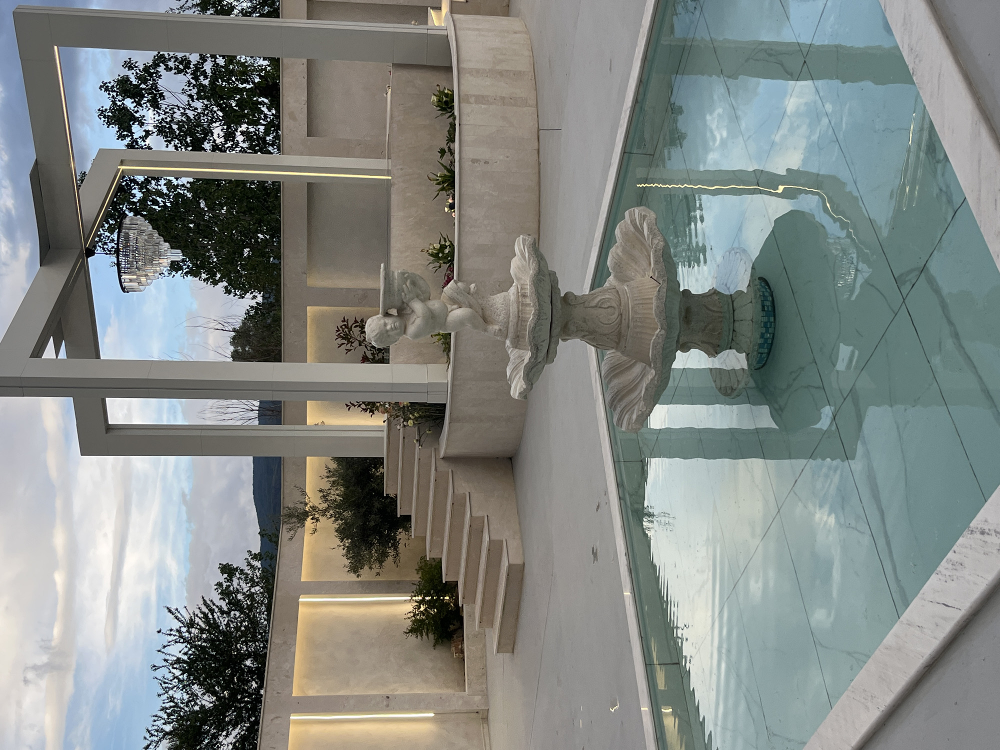 The image captures an outdoor scene featuring a decorative fountain. The fountain, made of white stone or marble, is located within a tiled pool of water. An angelic statue adorns the fountain. The scene is framed by a modern, white architectural structure with integrated linear lighting. In the background, tiered planters with greenery ascend a wall. In the far distance, a mountain range is visible under a partly cloudy sky. Based on the metadata, the location of the image is Gotse Delchev, Bulgaria.