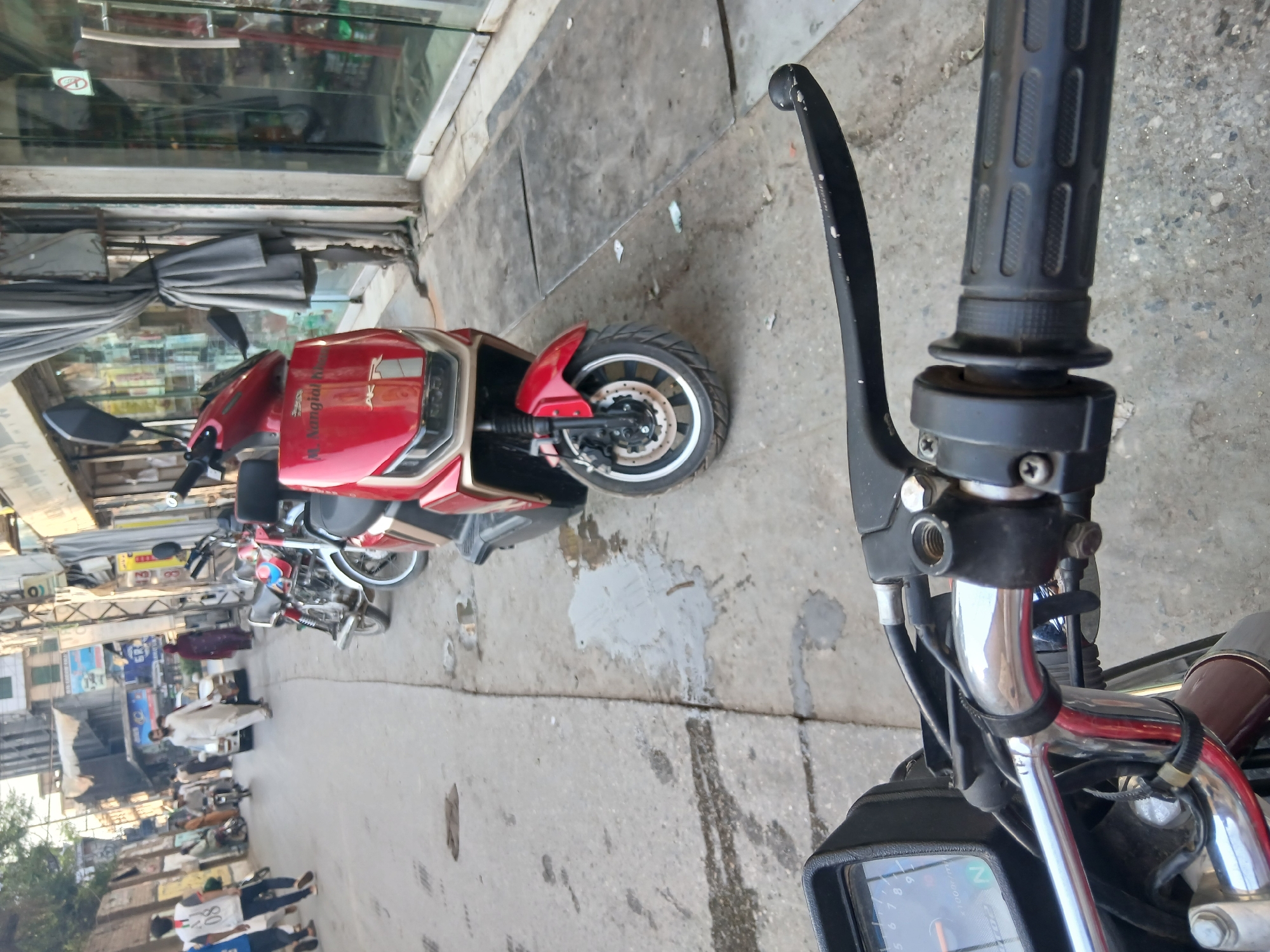 The image depicts a street scene in Peshawar City Tehsil, Pakistan, likely during daylight hours. The foreground is dominated by the handlebars and instrument cluster of a motorcycle, suggesting the photo was taken from the rider's perspective. The motorcycle has a silver handlebar, a black grip, and a partially visible instrument panel with a needle indicator.
In the midground, several vehicles are present. A prominent red scooter with gold accents is parked on the right side of the street. To its left, two other motorcycles, one red and white and another red, are also parked. Further down the street, a white car is visible, and several pedestrians are walking. One man in a white shalwar kameez is walking towards the viewer, while others, including a woman in a dark headscarf, are also visible. The street itself is paved and appears somewhat worn.
The background shows buildings lining the street, with awnings and shops visible. The architecture suggests an urban environment. The overall impression is a typical street scene in a Pakistani city, with a mix of vehicles and pedestrians navigating the thoroughfare.