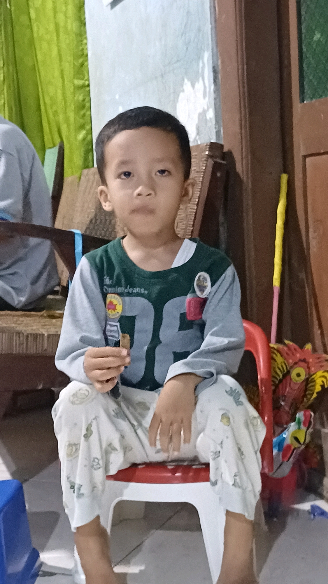 A young boy is seated on a white plastic chair with a red seat. He is wearing a long-sleeved shirt with a dark green collar and sleeves, and light grey body. The shirt has "the comic jeans." and "96" printed on it. He is also wearing light-colored pants with a pattern of small green figures. The boy is holding a thin, dark object in his right hand. To the right of the boy, a brightly colored dragon-like mask is visible, along with a yellow and pink pole. The background includes a wooden door frame and what appears to be green curtains. The overall setting suggests an indoor environment in a residential area.