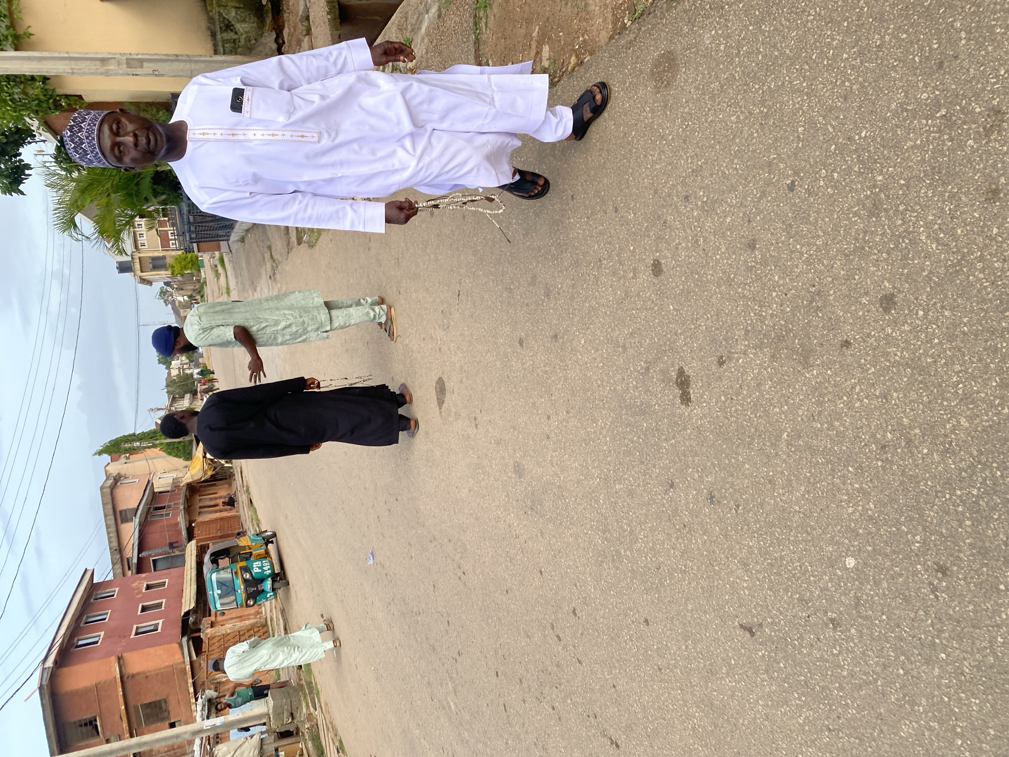 In Jos, Nigeria, a street scene unfolds with a mix of daily life and cultural attire. In the foreground, a man in a white kaftan and a patterned cap walks towards the viewer, holding a string of prayer beads. Behind him, another man in a black kaftan is captured in motion, and further back, a person in a light green kaftan stands facing away from the camera. A vibrant green and yellow auto-rickshaw is parked on the side of the road, adding a splash of color to the scene. The background reveals a row of buildings with orange and earth-toned facades, characteristic of the local architecture. The street is paved with asphalt, showing signs of wear and tear, with some debris scattered on the surface. The sky above is overcast, suggesting a cloudy day. The overall atmosphere conveys a sense of an ordinary day in a Nigerian urban setting.