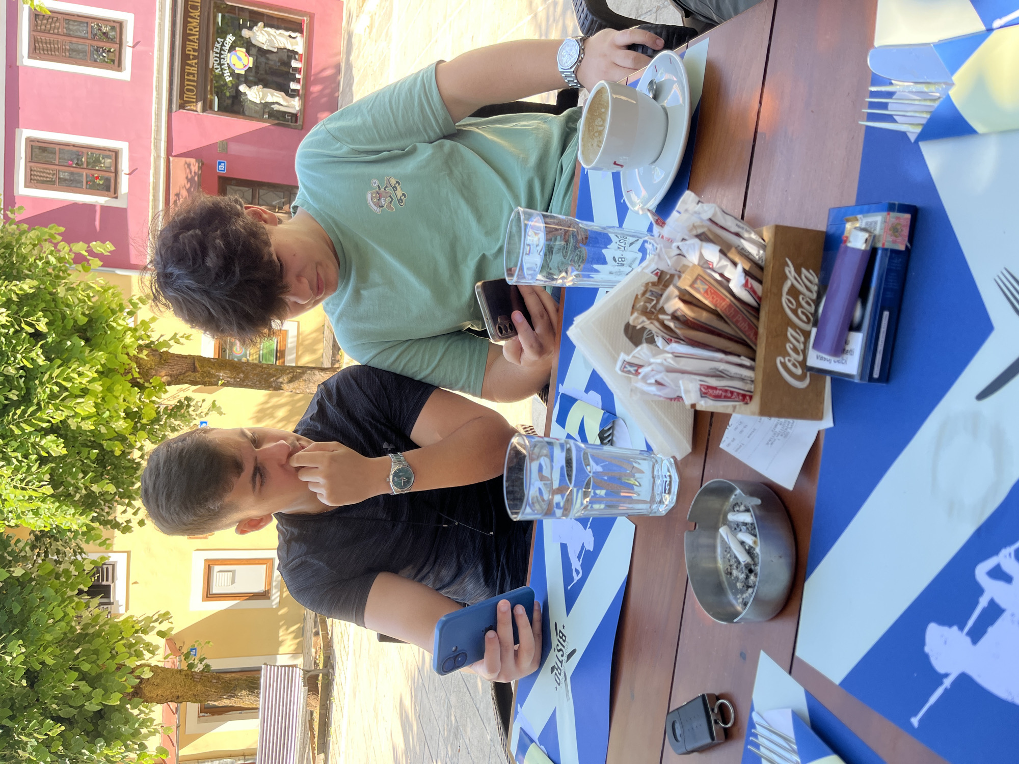 Two young men are seated at a wooden table outdoors, possibly at a cafe on Njegoševa Street in Cetinje, Montenegro. The man on the left, wearing a black t-shirt, is looking down at his blue smartphone, which he holds in his left hand. He has a silver watch on his left wrist and his right hand is touching his chin. The man on the right, wearing a light green t-shirt with a cartoon graphic, is also looking down at a smartphone in his right hand. He has a silver watch on his left wrist.
On the table, in front of the man on the left, is a glass of water, and to his right, a blue and white patterned placemat. In front of the man on the right is another glass of water, a white cup and saucer with remnants of coffee, and a wooden box filled with sugar packets and other small sachets, with "Coca-Cola" printed on the side. A pack of cigarettes with a purple lighter on top is also on the table. To the left of the table, there is an ashtray with cigarette butts and a set of car keys. The background shows buildings with colorful facades, green trees, and dappled sunlight.