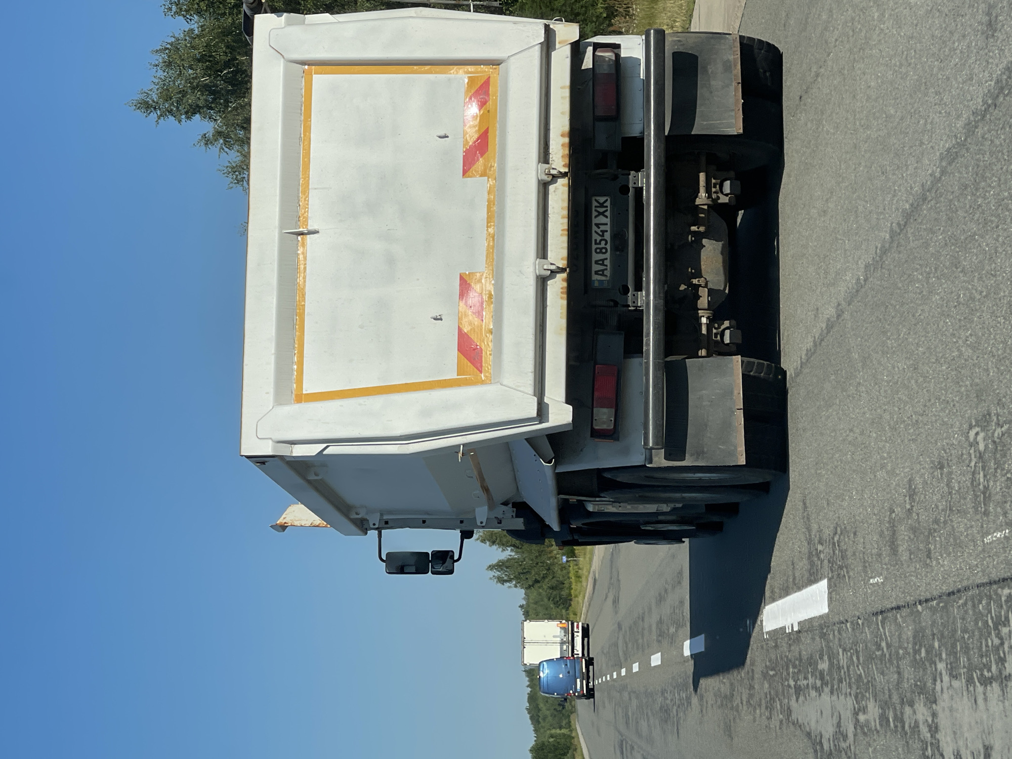 The image depicts a sunny day on a two-lane highway in Pyrizhky, Ukraine, with a clear blue sky and trees lining the right side of the road. The foreground is dominated by the rear of a white dump truck with a large, mostly white cargo bed. The truck has a license plate reading "AA 8541 XK". Two red and yellow reflective stripes are visible on the lower part of the truck's cargo bed, and there are some rust stains around the hinges. The truck is in the right lane of the highway. In the left lane, further ahead, is a blue van, followed by a white semi-trailer truck. The highway has white lane markings.