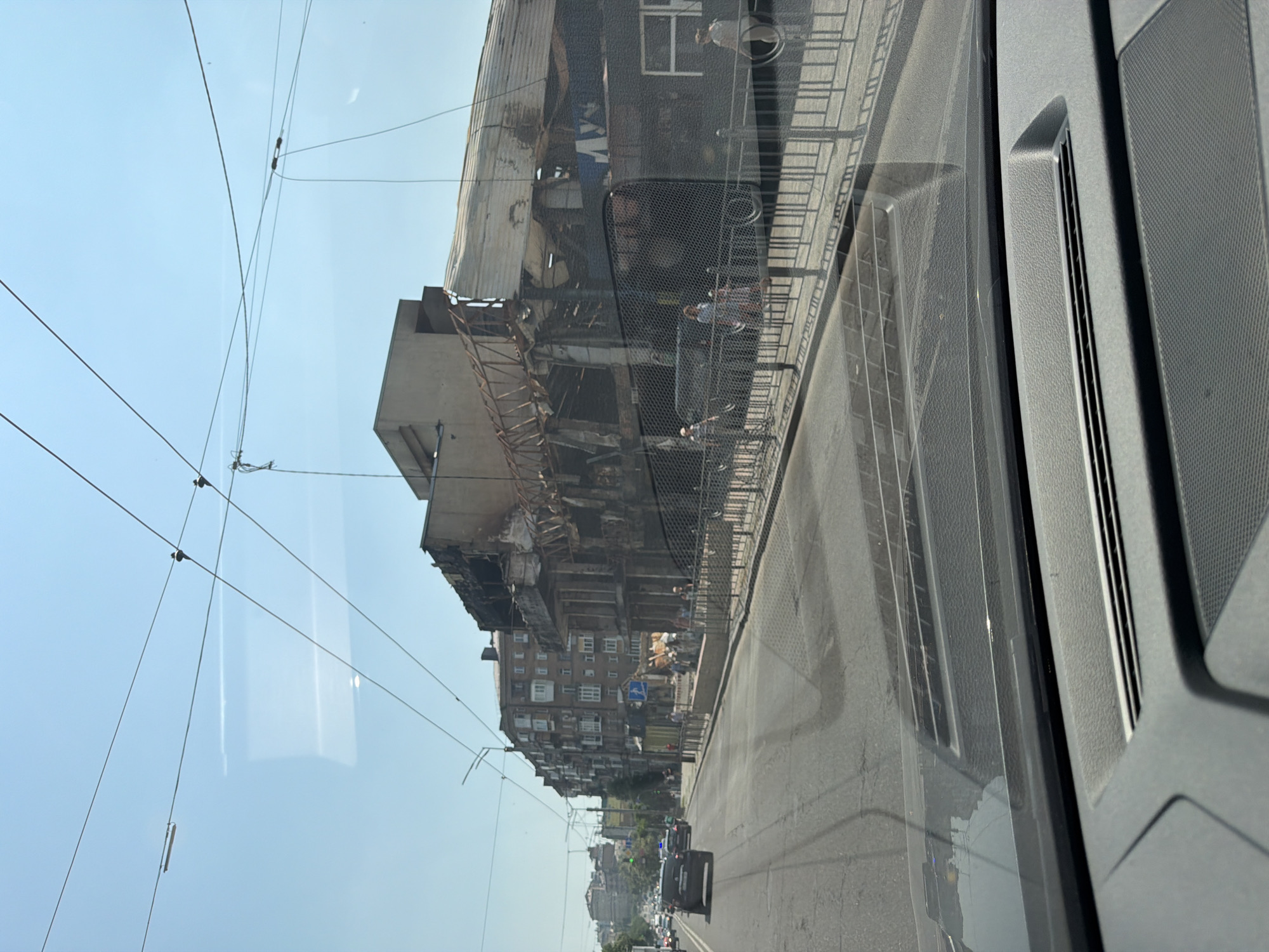 This is a photograph taken from a vehicle, showing a street scene in Kyiv, Ukraine. The right side of the image features a severely damaged building with its roof caved in and structural elements exposed. The facade of the building appears blackened and scorched, indicative of a fire. A metal fence runs along the sidewalk in front of the building. Several people are visible walking on the sidewalk. On the left side of the image, a street with several cars driving on it is depicted. In the background, there are apartment buildings and a clear blue sky with overhead wires. The photo captures a daytime scene.