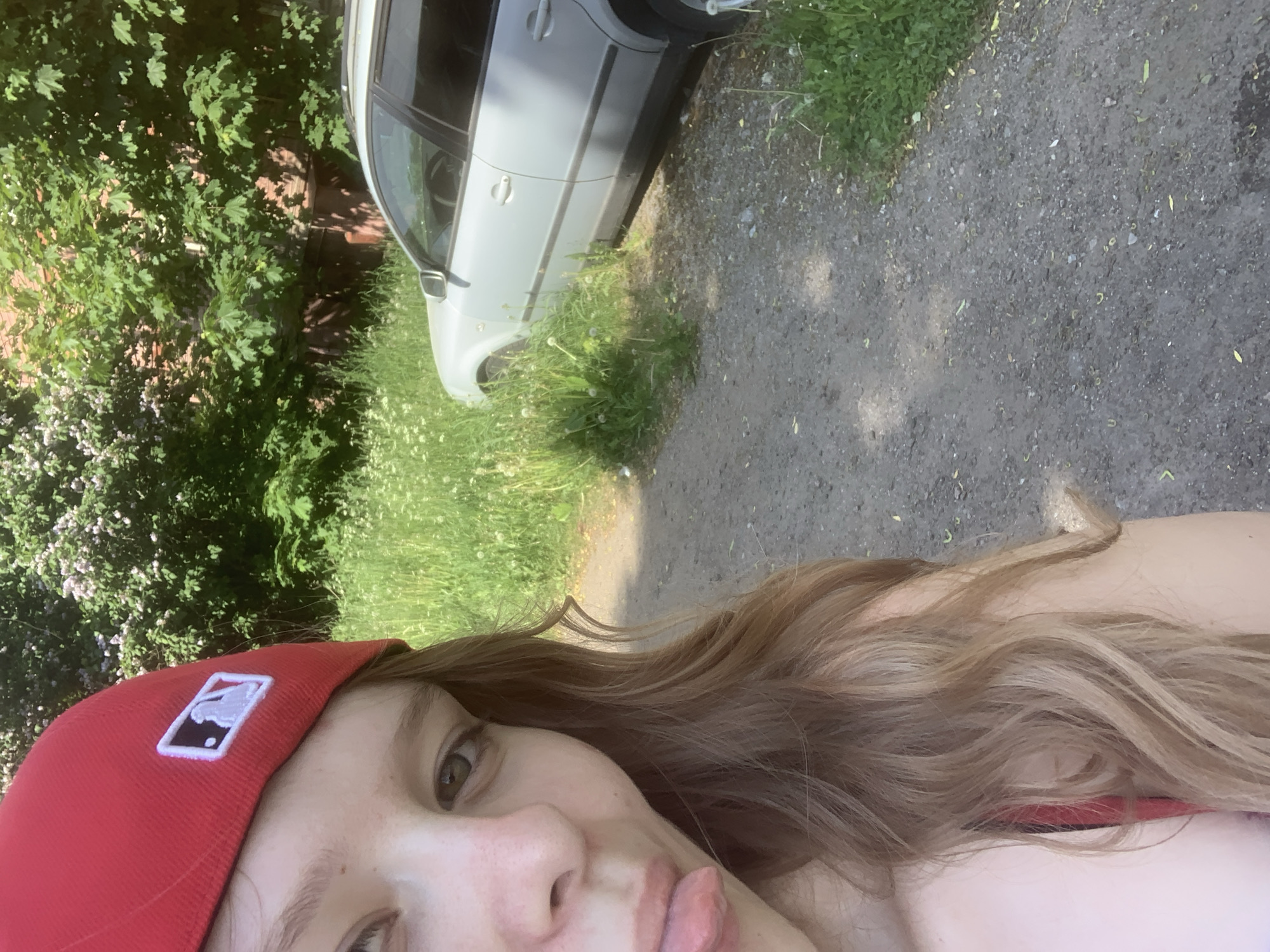 A young individual, partially visible on the left side of the frame, is taking a selfie in Priozersk, Russia. They are wearing a red baseball cap adorned with a white embroidered Major League Baseball (MLB) logo. Their left eye, nose, and mouth are visible, with the tongue extended playfully. Long, wavy, light brown hair falls over their shoulder. The background features an unpaved dirt and gravel path on the lower right, bordered by lush green grass and weeds, some bearing white blossoms. A light-colored sedan or station wagon is parked amidst the tall grass on the right. Dense green foliage from trees and bushes, some with light-colored flowers, occupies the upper right, partially obscuring a reddish-brown brick building. The scene is illuminated by strong sunlight, indicating an outdoor setting.