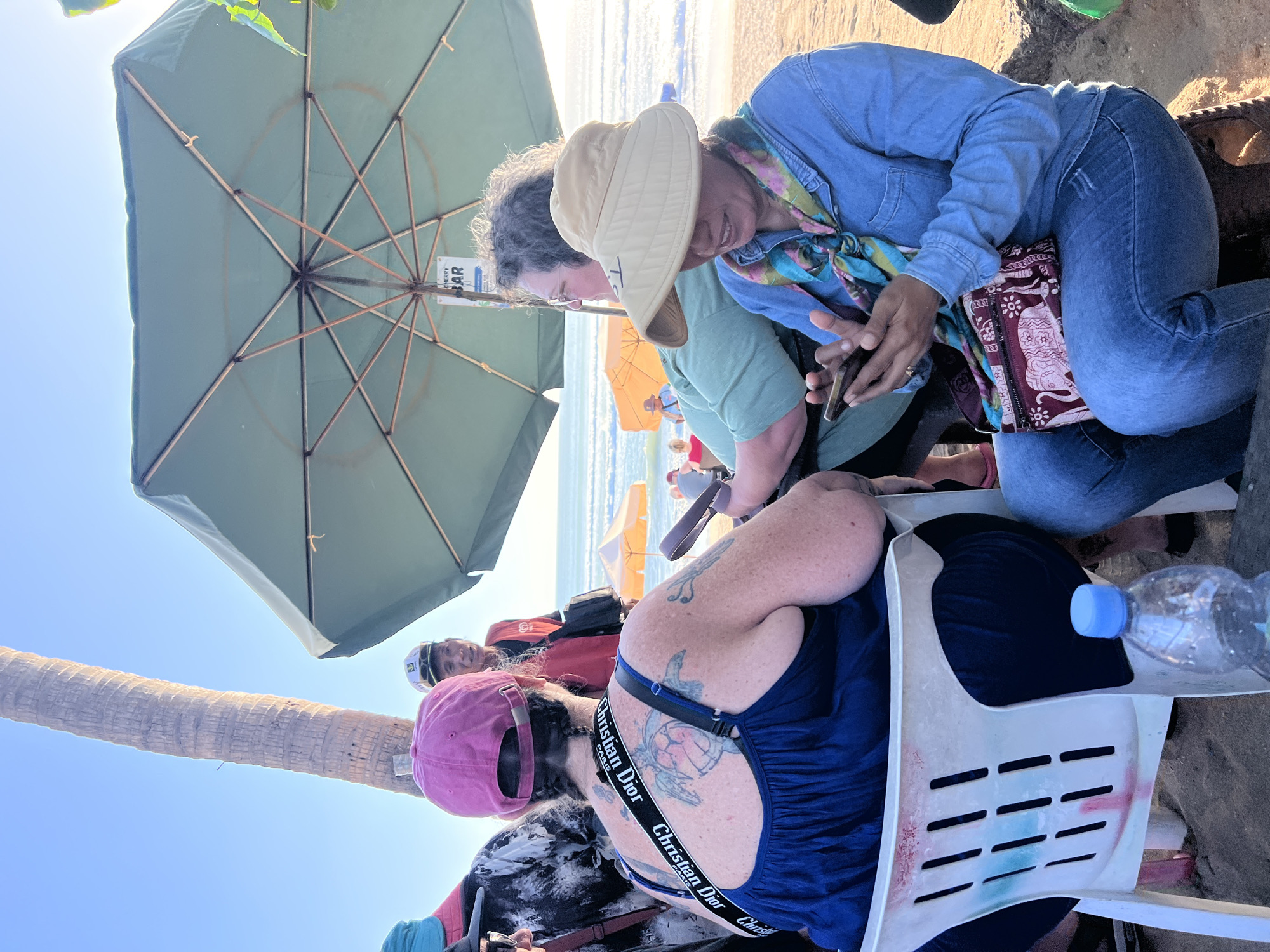 At a beachfront cafe in Kuta, Indonesia, four people are seated under a large green umbrella. The woman in the foreground, wearing a wide-brimmed hat and denim shirt, is engrossed in her smartphone. To her left, another woman with a pink hat and a blue dress is turned away from the camera, revealing a large tattoo of a turtle on her back. A man in a red vest and a white cap stands behind her, looking towards the sea. The fourth individual, partially visible, is wearing a green shirt. The sandy beach stretches out behind them, with the ocean and a few yellow umbrellas visible in the distance under a clear blue sky.