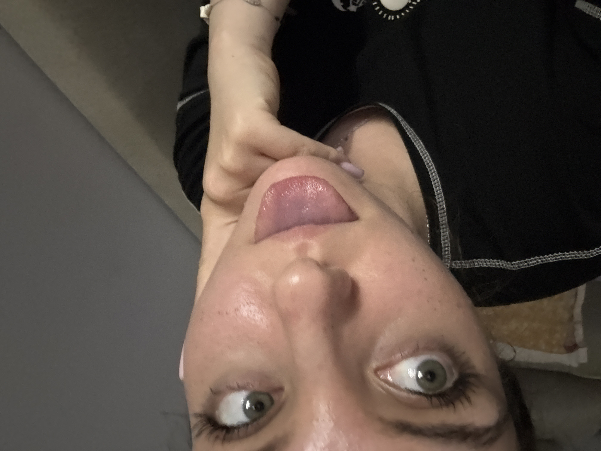 A selfie-style image shows a person, likely a female, with a close-up view of their face. The person is positioned upside down relative to the frame. Their eyes are wide open, and they are sticking out their tongue. The tongue appears to have a purplish tint. The person is wearing a black top with white stitching. The background includes a gray wall and a glimpse of what seems to be a cushion or textile with a yellow and white pattern. The location in Sliven, Bulgaria is unknown.