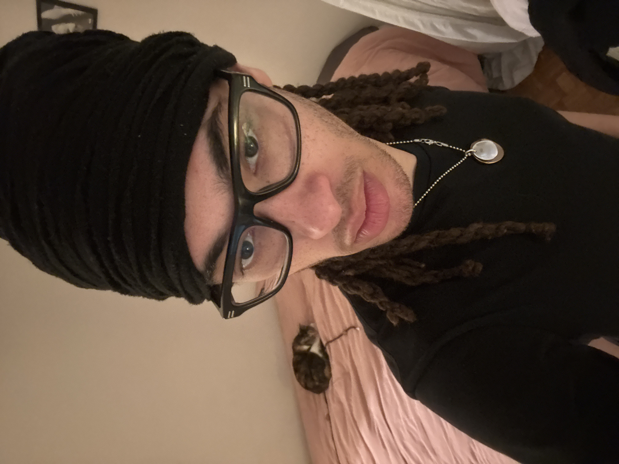 An adult individual is centered in a close-up, indoor selfie. They have a dark complexion, wear a black textured head covering, and rectangular black-framed glasses. Dark dreadlocks are visible at the sides of their face. They are wearing a black short-sleeved top and a silver ball-chain necklace with a two-layered circular pendant. Their gaze is directed towards the camera. In the background, to the left of the individual, a domestic cat with a calico/tabby fur pattern (brown, white, and dark patches) is curled up and resting on a bed with light pink/peach sheets. The wall behind the bed is a plain light color, and a dark-framed picture is partially visible on the upper right wall. The lower right edge of the image shows a segment of a light-colored wooden floor and white fabric. The image provides no specific visual information to determine a precise location within the city of Paris, France.
