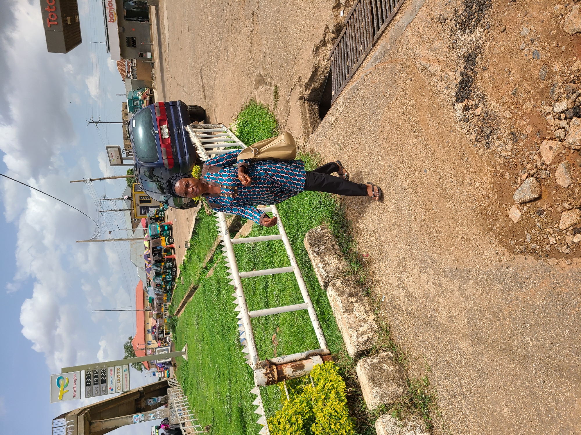 The image displays an outdoor scene in Gura Top, Nigeria, under bright daylight with a partly cloudy sky. A woman is positioned on the right side of the frame, walking or standing on a paved area bordering a grassy median. She is dressed in a long-sleeved top featuring a blue, green, and pink geometric pattern, black trousers, and open-toed sandals. She carries a light beige shoulder bag and has a dark head covering; her facial expression is neutral.
Behind the woman, a white metal barrier with spiked tops separates the pedestrian area from a strip of green grass. Further into the background, a dark blue car is parked, and a line of auto-rickshaws, commonly known as Keke Napep, can be seen along the street. The setting includes a TotalEnergies gas station with a prominent sign displaying fuel prices: "SUPER 197.5", "DIESEL 197.5", and "KEROSENE 220.0", alongside the brand name "QUARTZ". A "bonjour" convenience store sign is also visible as part of the station. Overhead, power poles and lines are present, and a concrete overpass or bridge is partially visible on the far left. The ground in the foreground shows a mix of asphalt and gravel, indicating some wear, with a metal grate covering a drainage channel. The scene captures typical urban infrastructure and daily activity in the region.