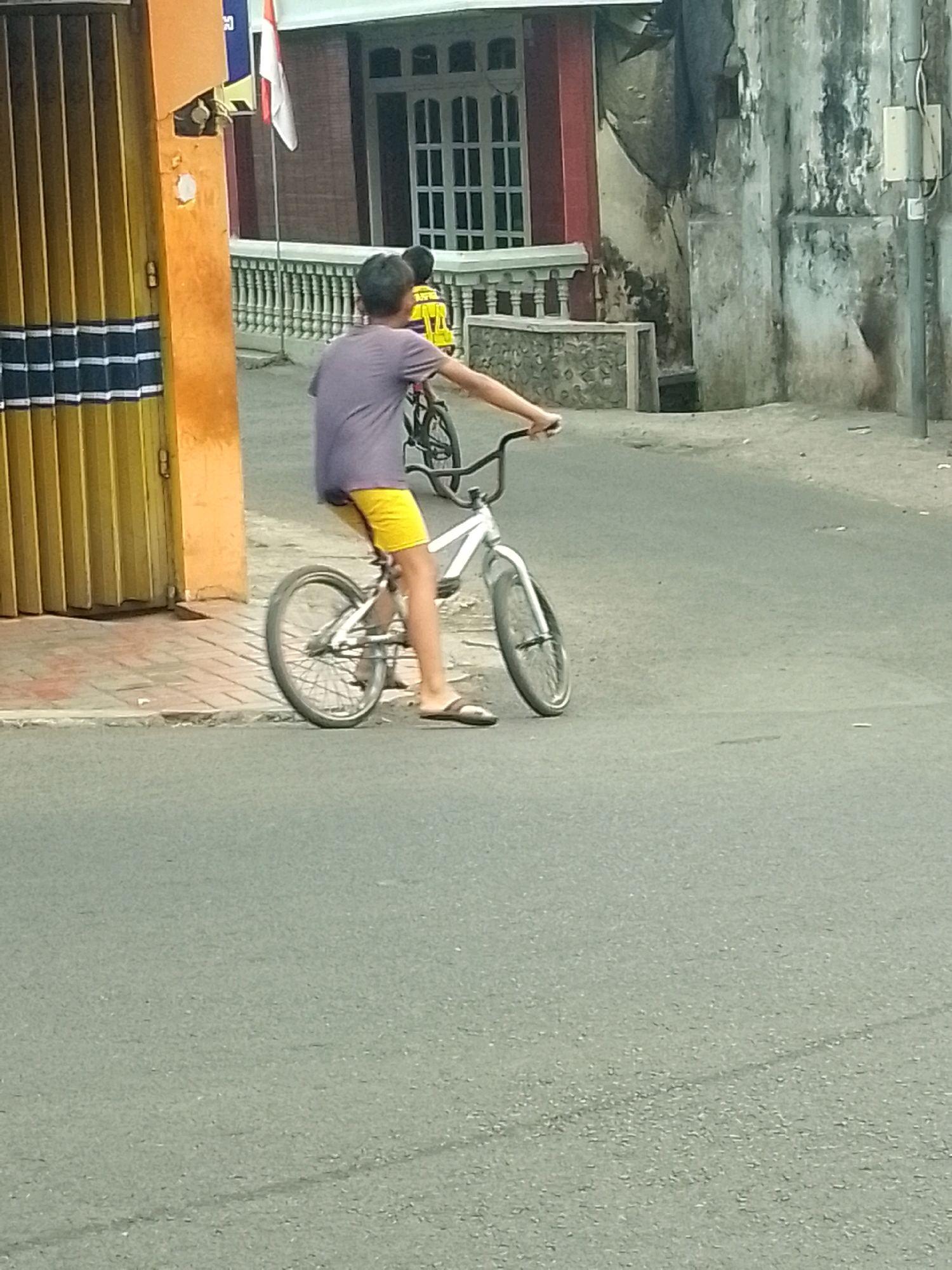 A young person with short, dark hair is riding a BMX bicycle on a paved road. They are wearing a purple t-shirt and bright yellow shorts. The bicycle has a white frame, black handlebars, and silver wheels. The rider's left foot is on the pedal and their right foot is on the ground, suggesting they are either starting or stopping. In the background, there is an orange building with a yellow corrugated metal gate and a white fence. There is also a glimpse of a building with a red facade and windows. The scene appears to be in an urban setting in Jember, Indonesia, evidenced by the architecture and the style of the buildings.