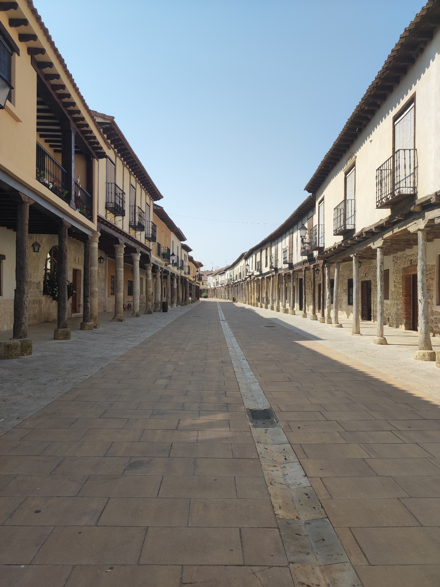 The image depicts a street scene in Ampudia, Spain, characterized by traditional architecture. On either side of a wide, paved walkway, two-story buildings line the street. These buildings feature a consistent architectural style: a ground floor with stone walls and wooden pillars supporting a covered walkway or arcade, and an upper floor with balconies adorned with black metal railings. The buildings are painted in light ochre and cream tones, with tiled roofs. The walkway is paved with rectangular stone tiles, and a central drainage channel runs down the middle of the street. The sky is clear and blue, indicating fair weather. The street is empty of people, suggesting it is early morning or late in the day. The overall impression is one of quiet, historical charm.