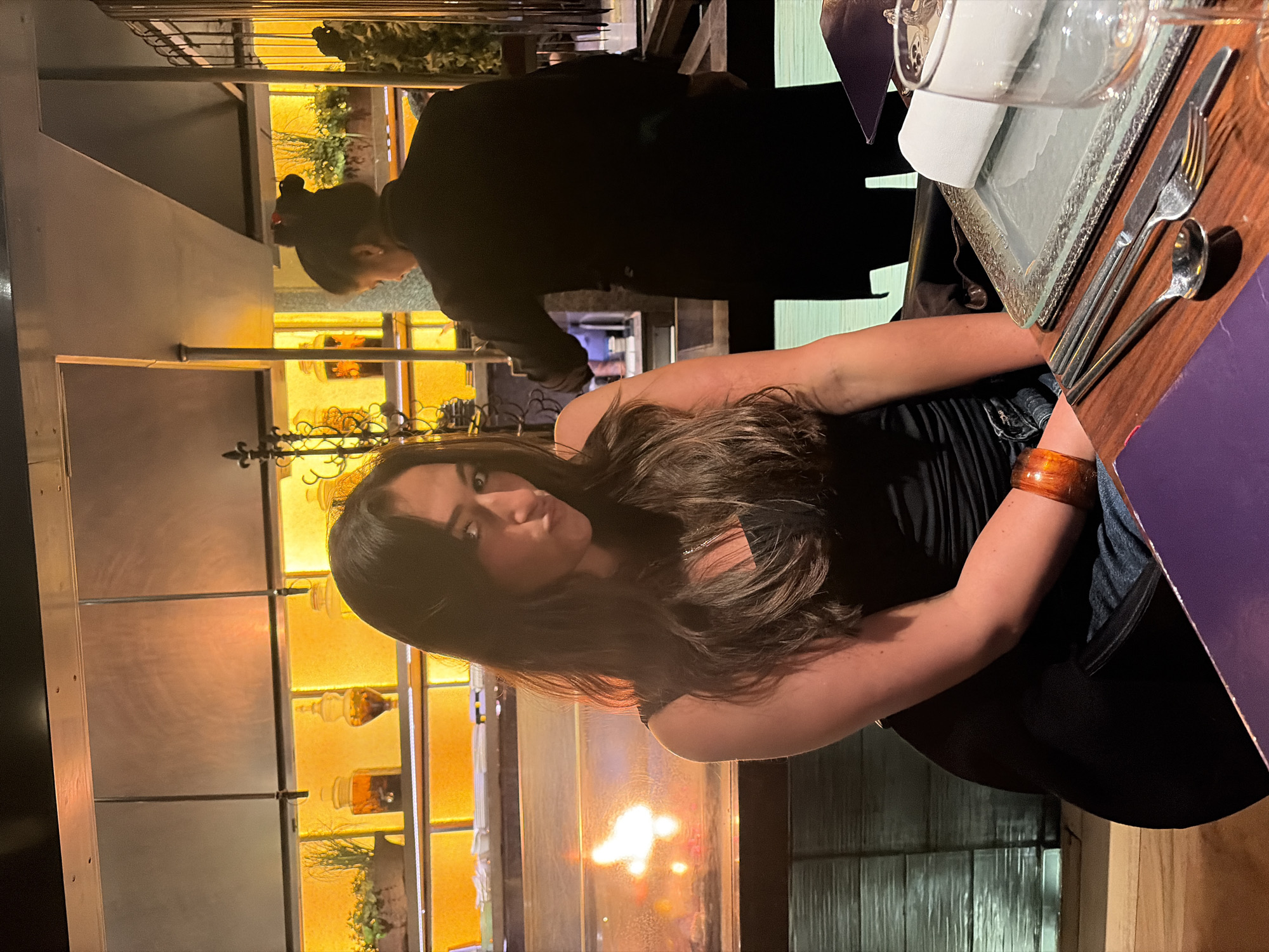 The image depicts a person with long brown hair sitting at a table in a restaurant. The person is wearing a black strapless top. The table has place settings including silverware and glassware. In the background, there is a second person wearing black standing. The restaurant has a warm, ambient lighting with what appears to be decorative bottles and jars on a shelf or display. Outside, the city is barely visible through a window but it is unknown.
