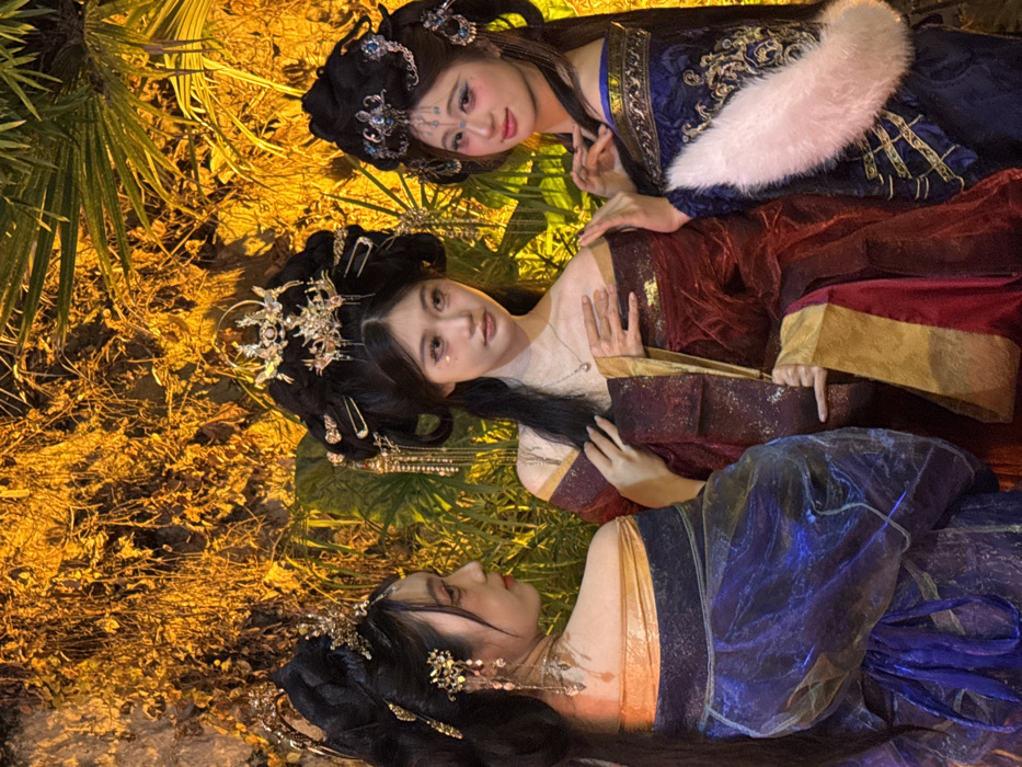 Three women in traditional Chinese attire are positioned horizontally, close together. Their elaborate black hairstyles are adorned with ornate, silver-toned headdresses. The woman at the top is wearing a blue and white fur-trimmed robe, the middle woman dons a dark red robe with gold accents, and the bottom woman is in sheer, shimmering blue attire. They are lying on what appears to be straw or dried grass, with foliage visible in the background. The warm lighting casts a golden hue overall.