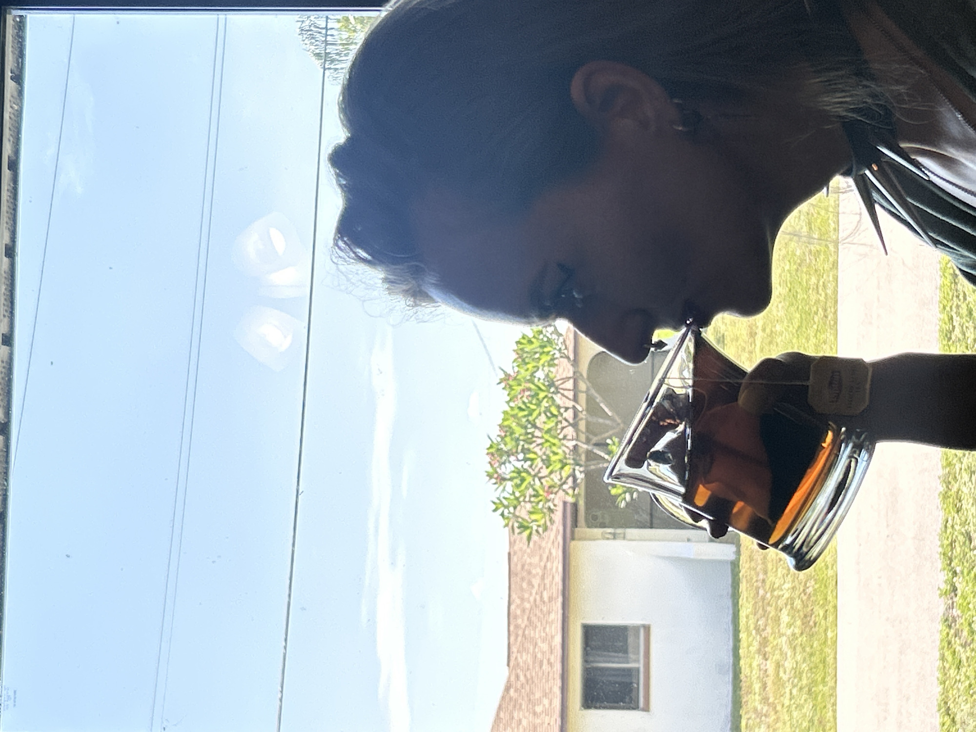 A woman is viewed in profile from the chest up, drinking from a glass of tea, with the scene set against a bright blue sky and a residential building. The woman has dark hair styled up with some strands loose, and she is wearing a dark, spiked choker and a dark necklace. She has a nose ring and an earring visible. The glass of tea is large and rectangular, with a tea bag tag hanging over the rim. Behind her, through a window, a white house with a brown tile roof is visible, along with a green tree with pink flowers. The overall lighting suggests it is daytime. The location is stated to be Cape Coral, Florida, United States of America, implying a sunny, suburban environment.