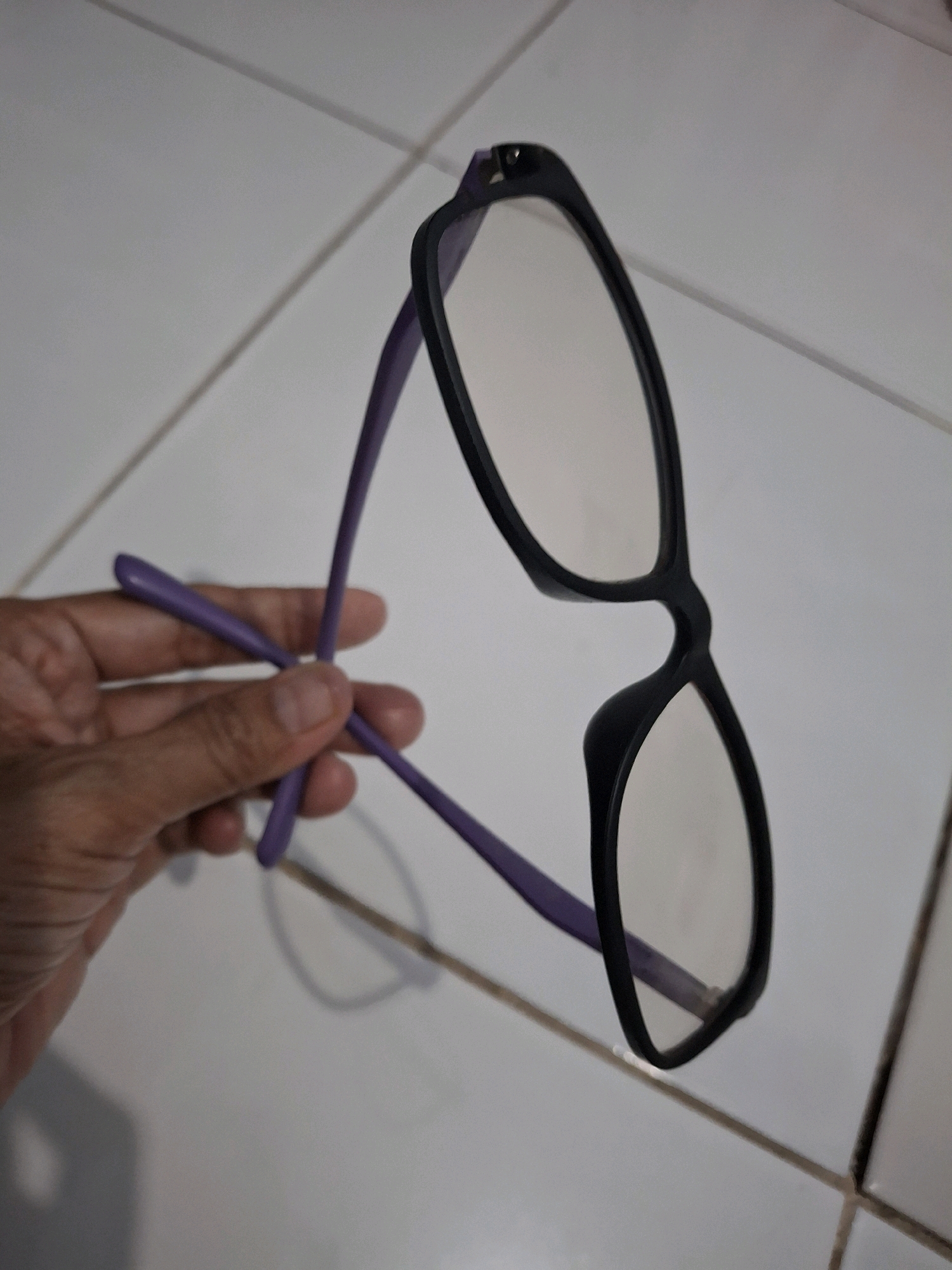 A pair of eyeglasses with black frames and purple arms is being held by a person's left hand, which is visible in the lower left quadrant of the image. The eyeglasses are positioned diagonally, with the lenses facing upwards and to the right. The background consists of a tiled surface, likely a floor, with visible grout lines. The tiles appear to be a light color, possibly white or beige. There are no other prominent objects or features in the scene. The lighting suggests an indoor environment. The image does not provide any specific information that would allow for identification of its location within Lumajang, Indonesia.