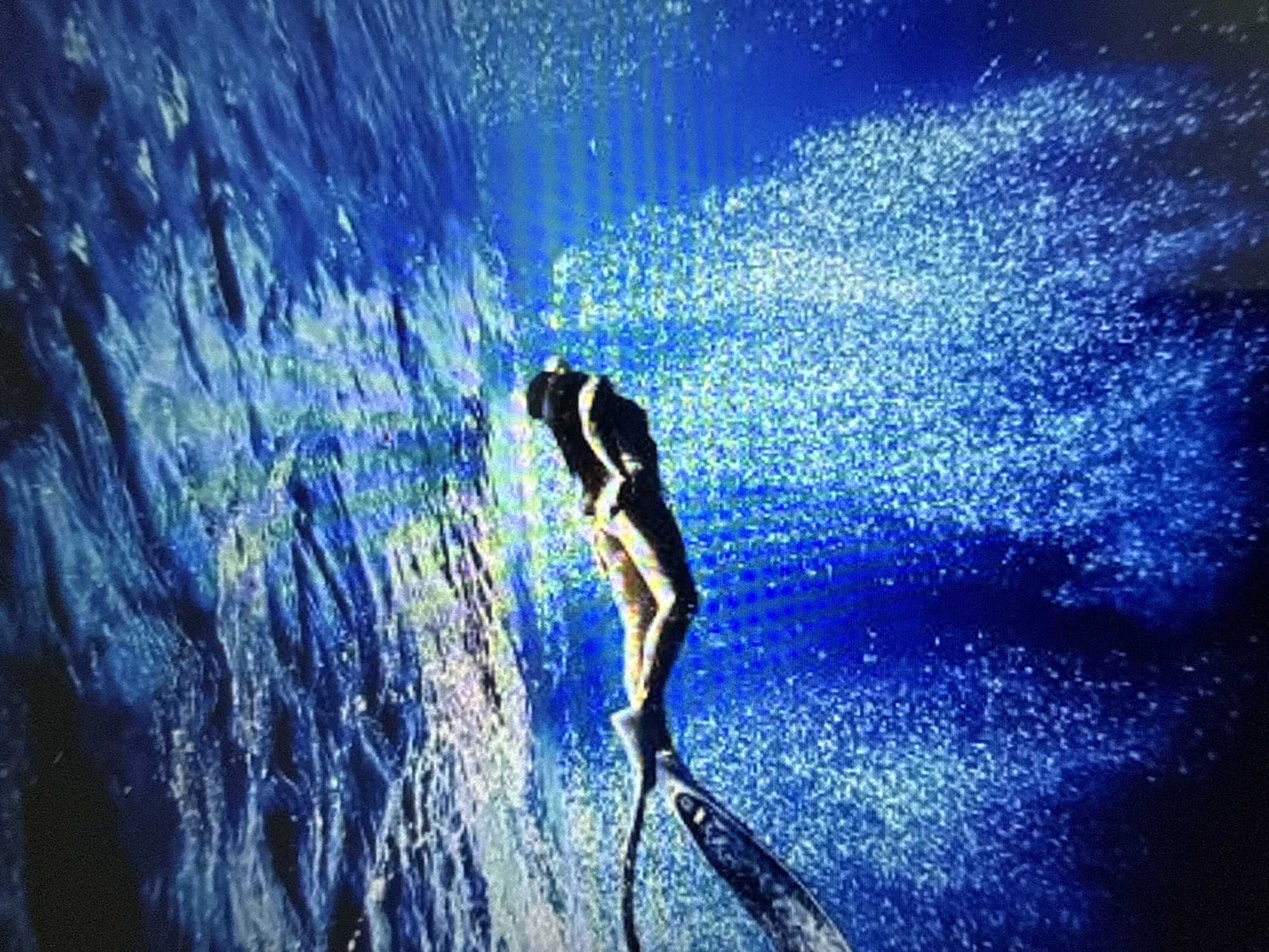 A person is swimming underwater, wearing a snorkel and mask, and a monofin on their feet. The water is a deep blue, and there are many bubbles surrounding the swimmer. The sunlight filters down from the surface, creating a shimmering effect on the water. The scene appears to be in the ocean or a large body of water.