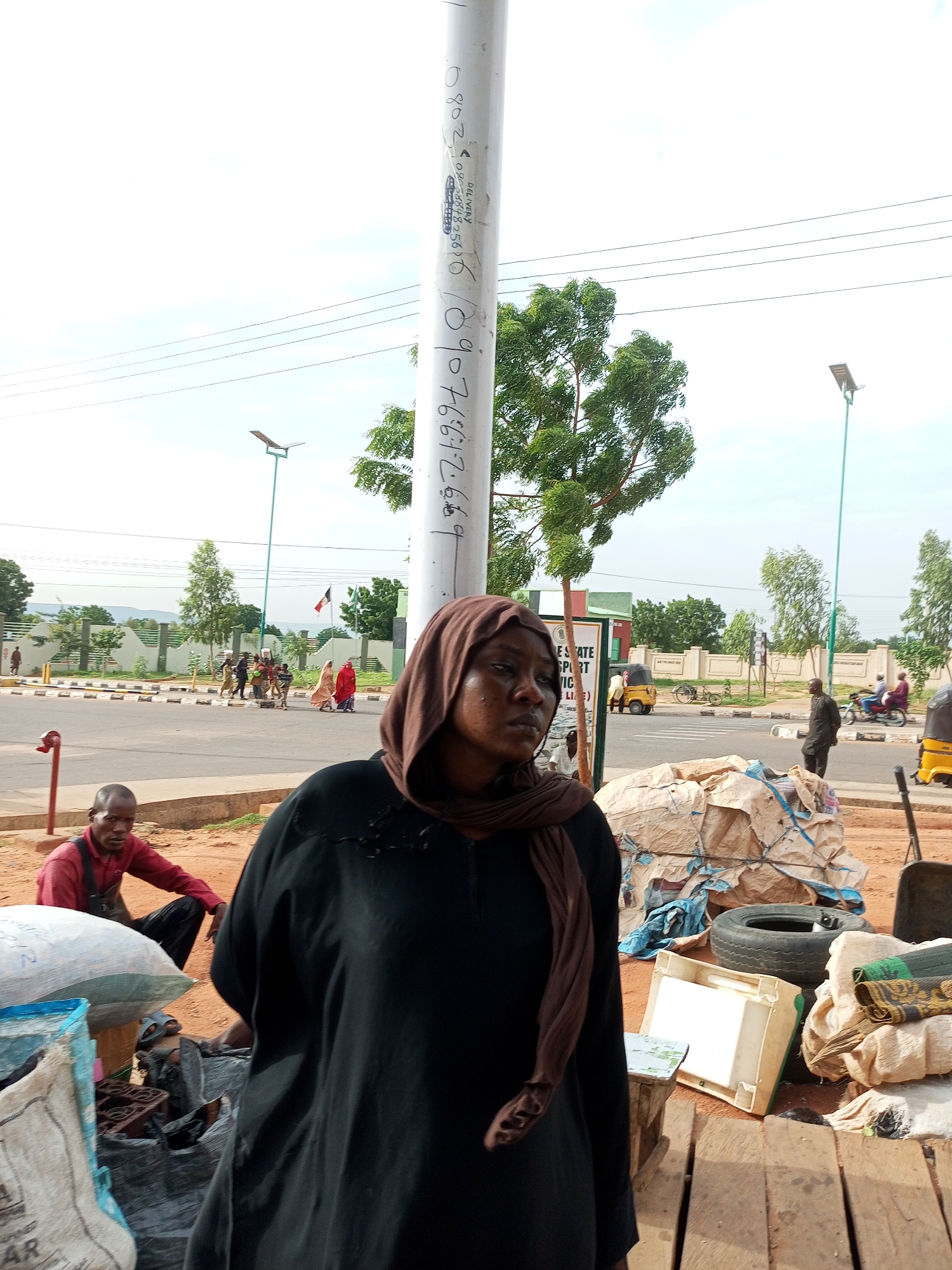 The image depicts an outdoor scene in Gombe, Nigeria. In the foreground, a woman wearing a dark abaya and a brown hijab is looking to the right. To her left, a man in a red shirt and dark overalls is seated. Piles of goods, including large sacks and what appear to be rolled-up materials, are situated in the foreground. In the mid-ground, a paved road is visible with several people walking, some carrying items on their heads. A fence lines the side of the road, beyond which are trees and buildings. A street light with a solar panel is prominent in the background, and electrical wires crisscross the sky. Traffic, including a yellow tricycle and motorcycles, can be seen on the road. The overall environment suggests a bustling street or market area.