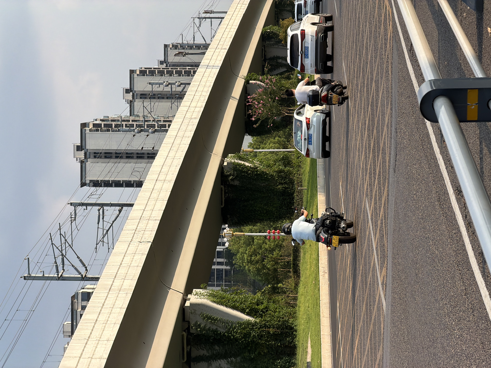 The image depicts a street scene in Jiangjin, China, under an elevated structure, likely for a railway or highway. A person is riding a dark-colored motorcycle with a yellow license plate that reads "XIA 803RR". The rider is wearing a light blue t-shirt and a black helmet. Following behind the motorcycle are several white vehicles, including two SUVs and a sedan. The vehicles are traveling on a paved road with yellow lane markings. In the background, several tall, modern apartment buildings are visible, as well as overhead power lines supported by poles. Green trees and ivy cover the pillars supporting the elevated structure, and a small tree with pink blossoms is situated in the middle ground. The sky is partly cloudy.