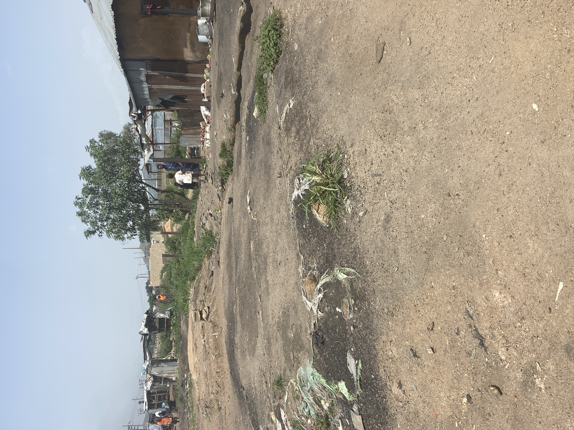 The image shows a street scene in Bukuru, Nigeria. The foreground is dominated by a dirt and concrete road with scattered debris, including plastic waste and some sparse vegetation growing through cracks. In the midground, several structures resembling informal housing with corrugated metal roofs are visible. Some people are standing near these structures. A large tree is present on the left side of the image, providing some shade. In the background, more buildings, possibly under construction with concrete blocks, and power lines are visible under a clear, light blue sky, suggesting it is daytime with fair weather. The overall impression is of a somewhat undeveloped urban or peri-urban environment. There is no visible text in the image.