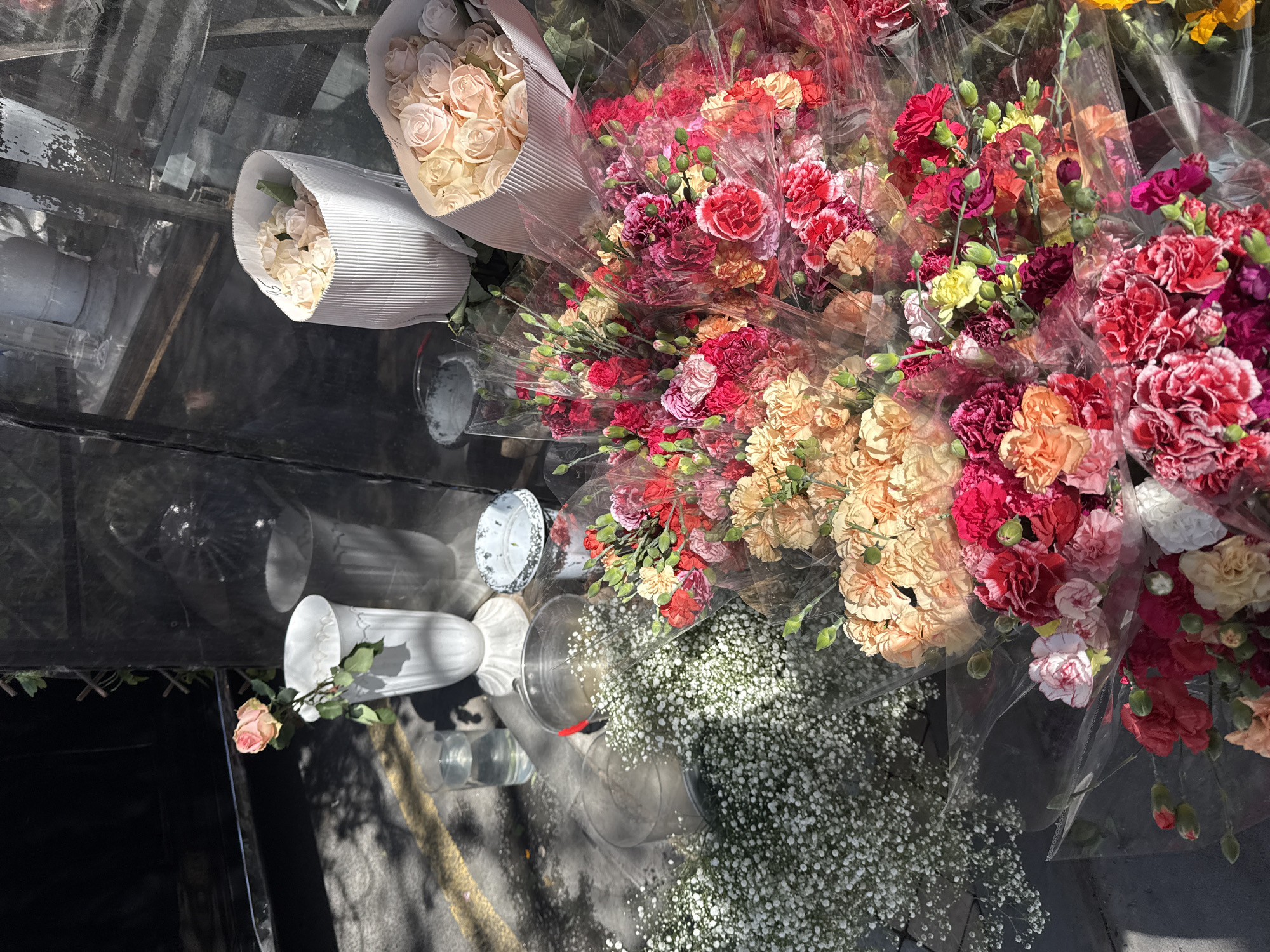 This overhead shot captures a vibrant display of fresh flowers, likely at a market stall or florist shop in Yerevan, Armenia. The flowers are arranged on a highly reflective, dark surface, possibly glass, which shows distorted reflections of the surroundings.
On the left side of the frame, two bouquets of light-colored roses, in shades of cream and pale pink, are neatly wrapped in ridged white paper. To their right, and filling a large portion of the image, are numerous bouquets of colorful carnations. These carnations are predominantly in rich hues of red, deep pink, fuchsia, orange, peach, and yellow, with some white accents, and many still have visible buds. Each carnation bouquet is individually wrapped in clear plastic.
Towards the bottom right, a large quantity of white baby's breath (Gypsophila) is clustered, also appearing to be wrapped or piled. On the reflective surface, several clear glass vases or containers are visible, some empty and one appearing to hold water. A white ceramic vase lies horizontally near the bottom left, with a single pale pink rose and some foliage resting beside it. Another white, possibly metallic, container with a speckled rim is also present.
The bright sunlight, indicated by harsh shadows and strong highlights, suggests a clear and sunny day. No discernible text is visible on the flower wrappings, vases, or other objects in the image. The scene conveys a sense of abundance and fresh floral beauty.