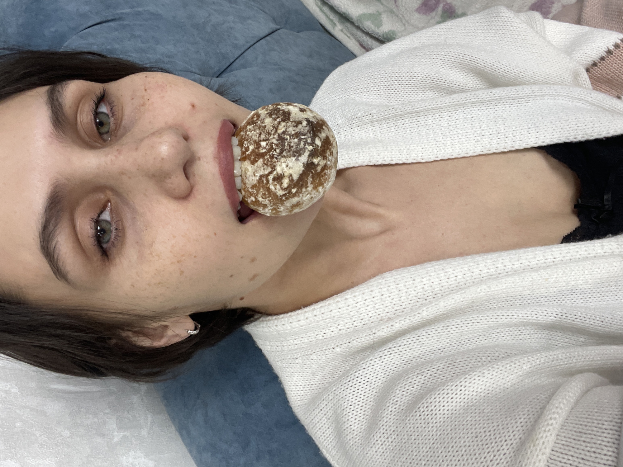 A young, light-skinned woman with freckles and green eyes is lying on a blue, possibly velvet, surface. She has short, dark hair and is wearing small hoop earrings. She is holding a round, brown and white object in her mouth, which appears to be a gingerbread cookie. She is wearing a white knit cardigan over a black top. The location is not explicitly stated in the image, and I cannot infer that the location is in the city of Izhevsk, Russia.