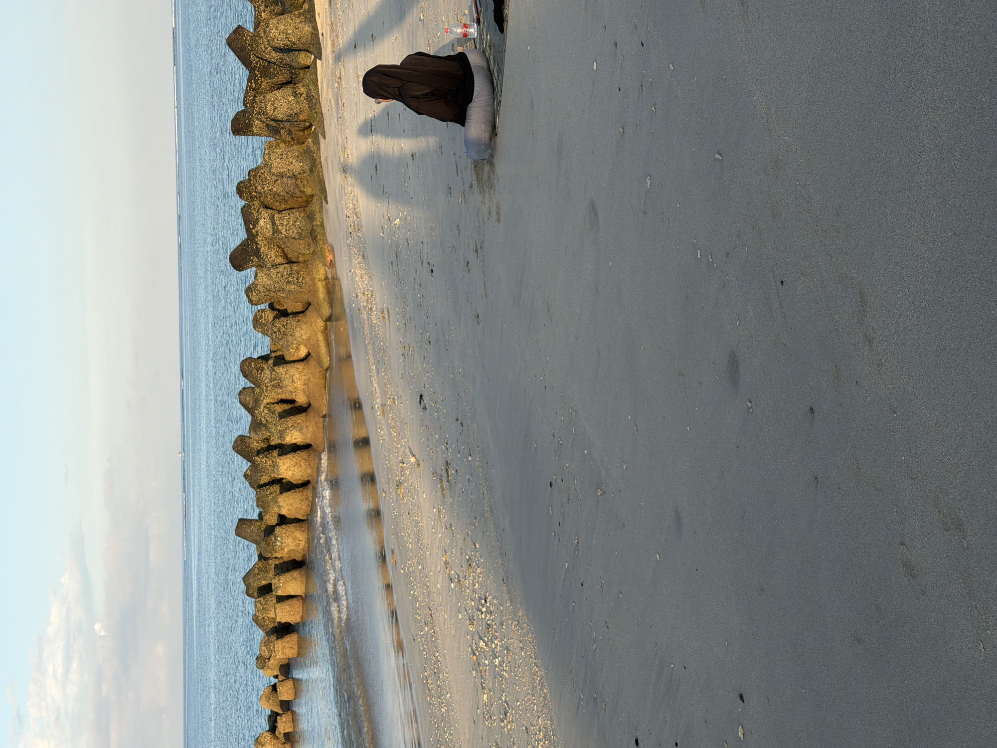 The image depicts a woman wearing a black hijab and light gray leggings sitting on a sandy beach next to a rock breakwater wall at Kuta Beach, Bali, Indonesia. The beach consists of dark gray sand with small shells scattered near the water line. A red and white plastic bottle sits beside her. The ocean is visible beyond the breakwater, with a light blue sky. The image is oriented vertically.
