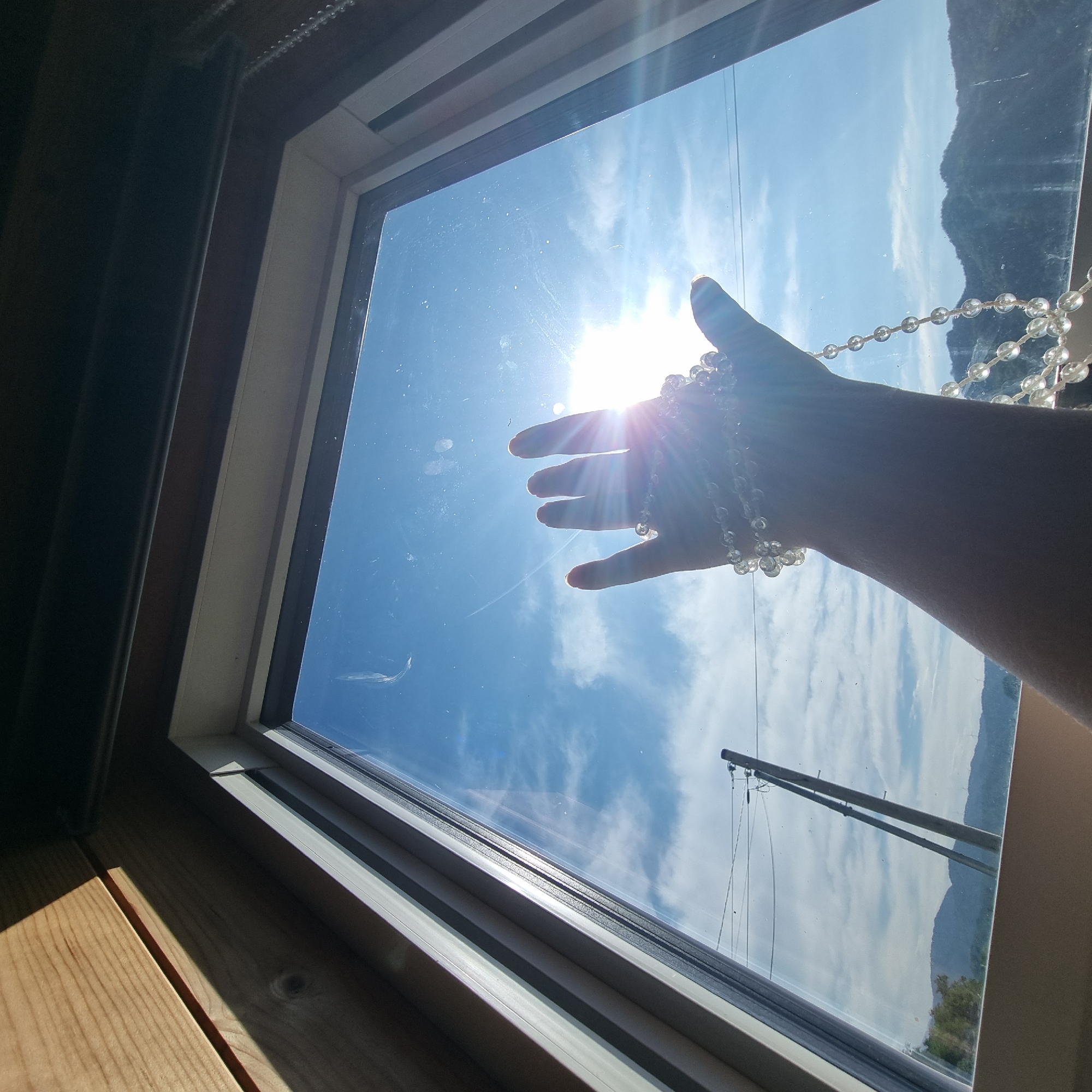 A person's hand, adorned with a string of clear, round beads, is held up towards a bright sun visible through a window. The sun is in the upper right portion of the frame, casting lens flare and sunbeams across the scene. The window looks out onto a bright blue sky with scattered white clouds. In the distance, silhouetted mountains can be seen, along with utility poles and power lines. The interior of the room, visible in the upper left of the frame, is made of wood paneling and is partially in shadow. The image appears to be taken from inside a building looking outward through a window. The location is identified as Dunnae-myeon, South Korea.