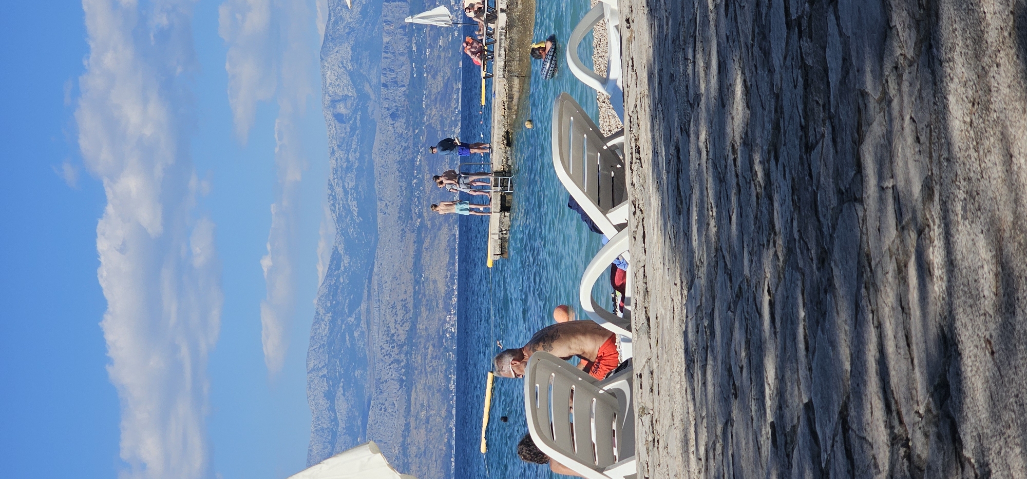 The image depicts a sunny day at a beach in Supetar, Croatia, with a focus on the coastline and the clear turquoise water. In the foreground, two white lounge chairs are visible on a stone-paved area, casting shadows. One chair is occupied by a man with tattoos on his back, wearing red swim trunks, and another person partially visible.
Further back, the sea is populated with people. A group of individuals stands on a small pier extending into the water, some appearing to be looking out at the sea. One person is in the water, lying on an inflatable ring. In the background, a mountain range with a town nestled at its base forms the horizon, under a bright blue sky with scattered white clouds. Yellow buoys are also present in the water.