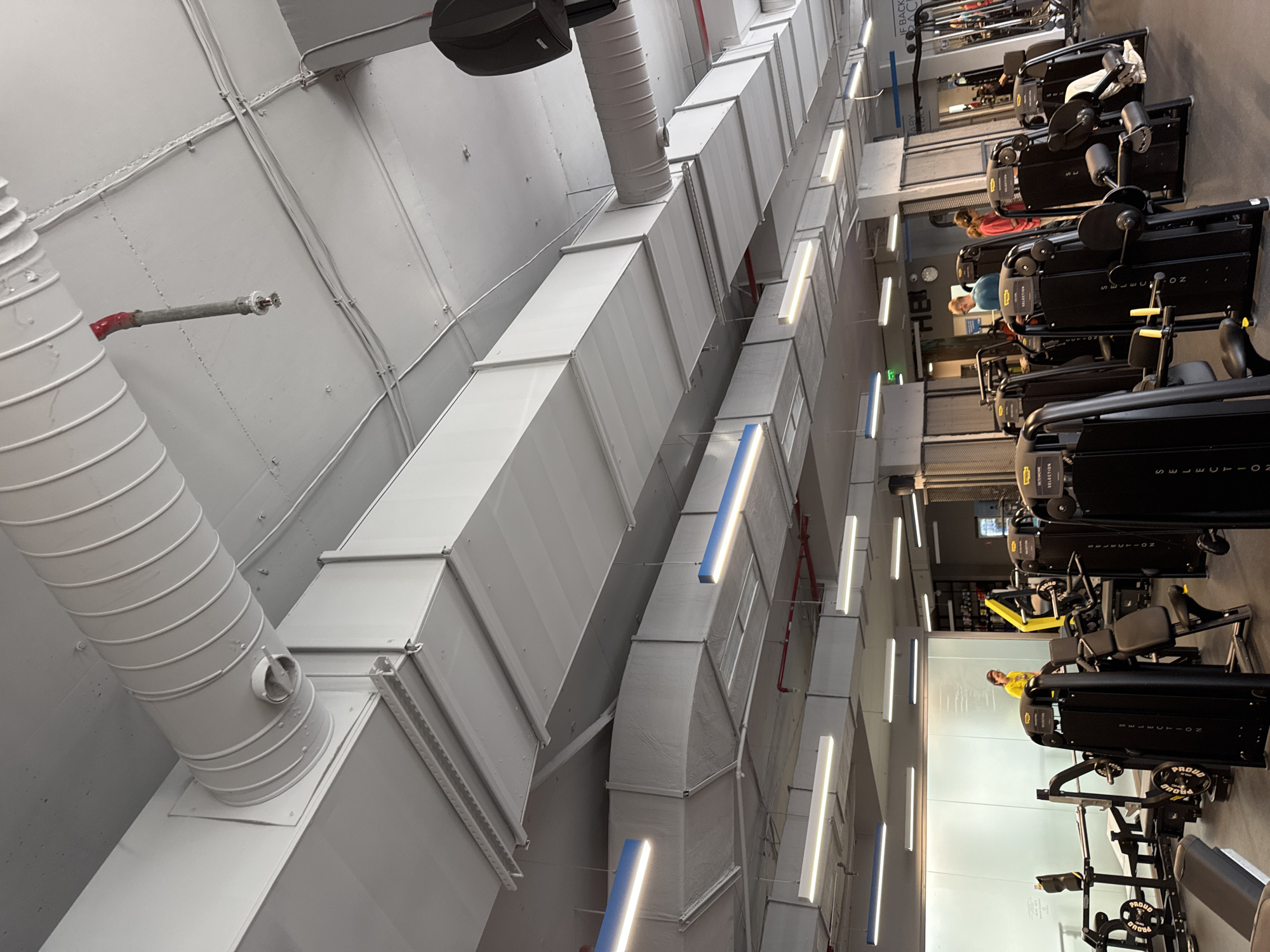 The image depicts the interior of a modern fitness gym, specifically identified as a facility belonging to the Pulse Fitness & Spa chain in Sofia, Bulgaria. The ceiling features extensively exposed white-painted infrastructure, including numerous parallel rectangular and spiral corrugated metal ventilation ducts, a visible red fire sprinkler head, and segments of red-painted fire suppression piping. Various linear LED light fixtures are suspended from the ceiling, some emitting white light and others featuring blue accents. Large black audio speakers are also mounted overhead. The floor is covered with dark, industrial-style gym matting. The gym is densely equipped with black 'Technogym Selection' series selectorized weight training machines, identifiable by branding, along with other strength equipment featuring yellow accents and free weight benches with 'PROUD' branded weight plates. Several individuals are present: one person in a yellow t-shirt stands near a frosted partition wall on the left, another in a blue t-shirt and glasses is situated in the mid-ground, and a third individual in a red long-sleeve shirt with headphones is visible on the right. Additional patrons are indistinctly visible further within the gym area among the equipment.