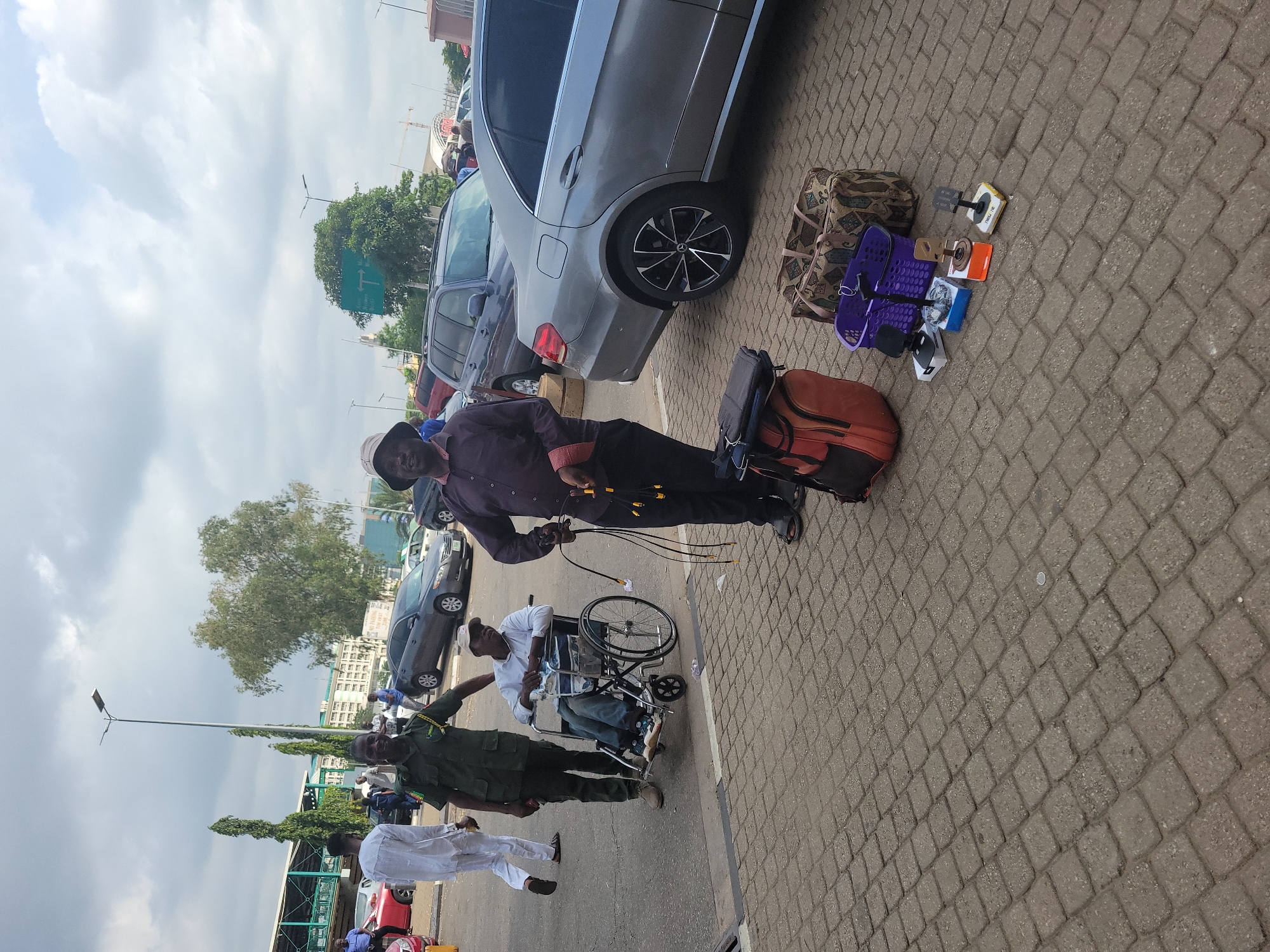 This outdoor image, set in Abuja, Nigeria, captures a daytime scene on a paved surface, likely a public thoroughfare, parking lot, or airport drop-off area. The sky above is cloudy but bright.
In the foreground, a man wearing a dark maroon shirt, black trousers, and a hat is standing, holding a bundle of electrical wires or cables. He is positioned next to a brown and black wheeled luggage bag. To his right, a grey sedan car is parked. On the ground beside the car, an assortment of items is displayed, including a patterned duffel bag, a purple plastic basket, and several small objects that appear to be foot pedals or electronic accessories.
Further back and to the left, a man in a white shirt and cap is seated in a manual wheelchair. A man in a green military-style uniform stands beside the wheelchair, seemingly interacting with or assisting the person in it. Another individual in a white traditional garment is visible behind them.
The background features more parked vehicles, tall street lamps, green trees, and distant buildings, suggesting an urban or semi-urban environment. The ground is covered with light brown interlocking brick pavement. The scene suggests a transient location where people and goods are moving. No discernible text is visible on any signs or objects.