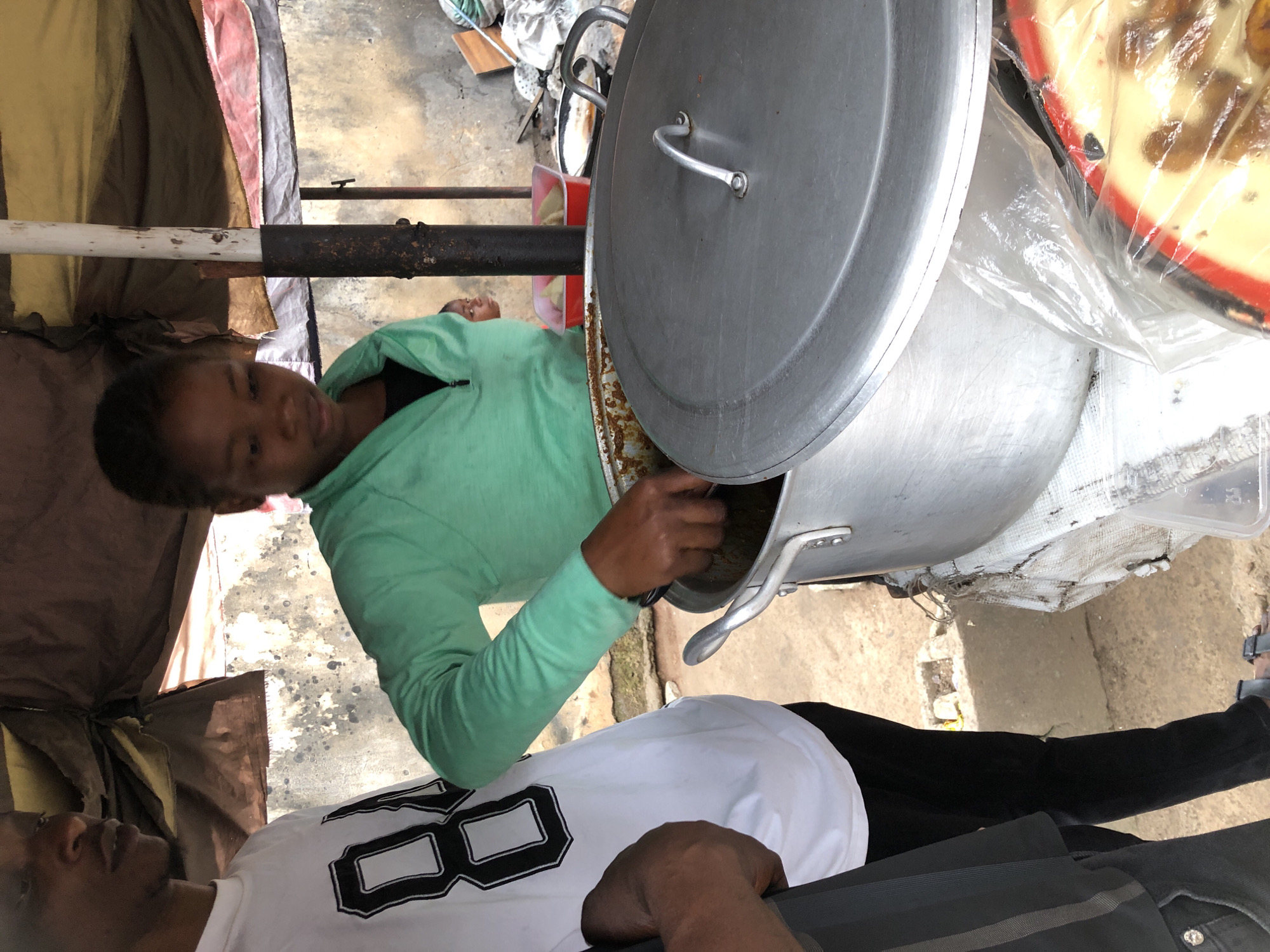 This image depicts an outdoor food stall in Palmgrove, Nigeria. A young woman wearing a green hooded jacket and black top is serving food from a large metal pot. She is using a utensil to stir the contents of the pot. Next to her, a person in a white t-shirt with a black graphic is partially visible, and another person is visible from the waist down in black pants and sandals. The stall is covered by a makeshift awning made of fabric and supported by a metal pole. In the background, there are concrete walls and other food items in red plastic containers and on plates. The overall scene suggests a busy street-side food vendor operation.