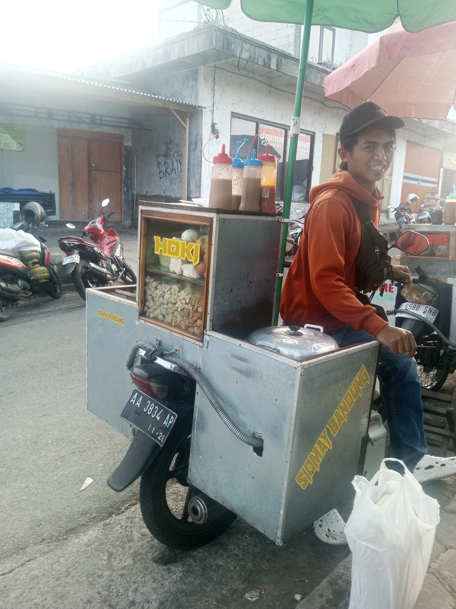 A young Indonesian man wearing an orange hoodie and blue jeans smiles at the camera from behind a food cart attached to a motorcycle. The food cart, which appears to be a mobile somay stand, is made of metal and displays the words "HOKI" and "SIOMAY BANDUNG" on its exterior. Inside a glass display case, various food items, likely somay and accompaniments, are visible. Three squeeze bottles containing sauces are also part of the cart's setup. The scene is set on a street in Kalibeber, Indonesia, with buildings and other motorcycles visible in the background. The man is seated on the motorcycle, with his hands resting on the food cart. A white plastic bag is placed on the ground next to the cart. The image is taken outdoors during daylight, with bright sunlight casting shadows.