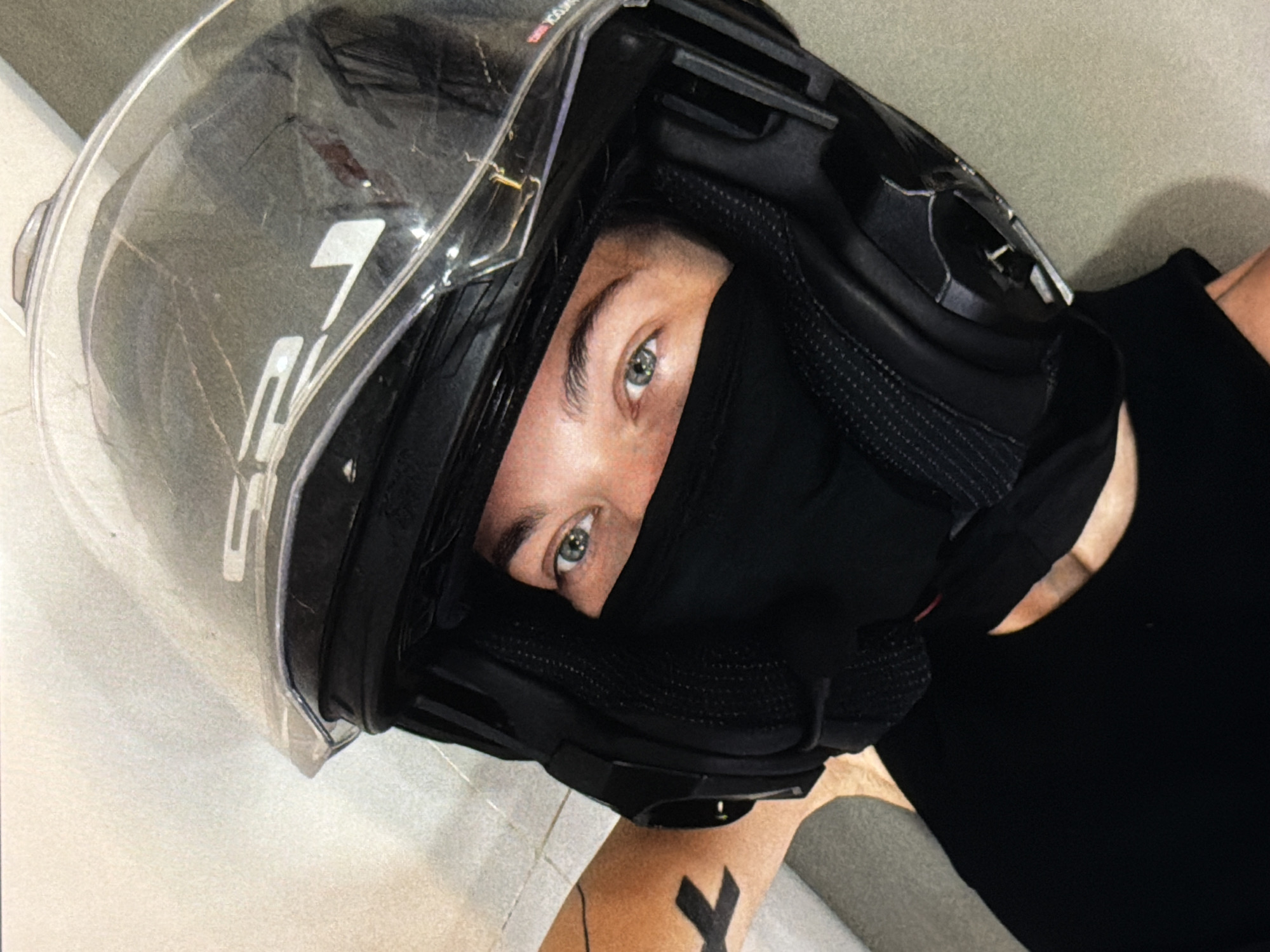 A person wearing a black balaclava and a motorcycle helmet is positioned at an angle against a light-colored background. The person's eyes are visible. A black tattoo is visible on the exposed part of the person's arm. The helmet is black with a clear visor and the logo "LS2" is visible on the visor. The image is a close-up shot. There is no indication of the location being Đà Nẵng, Vietnam.