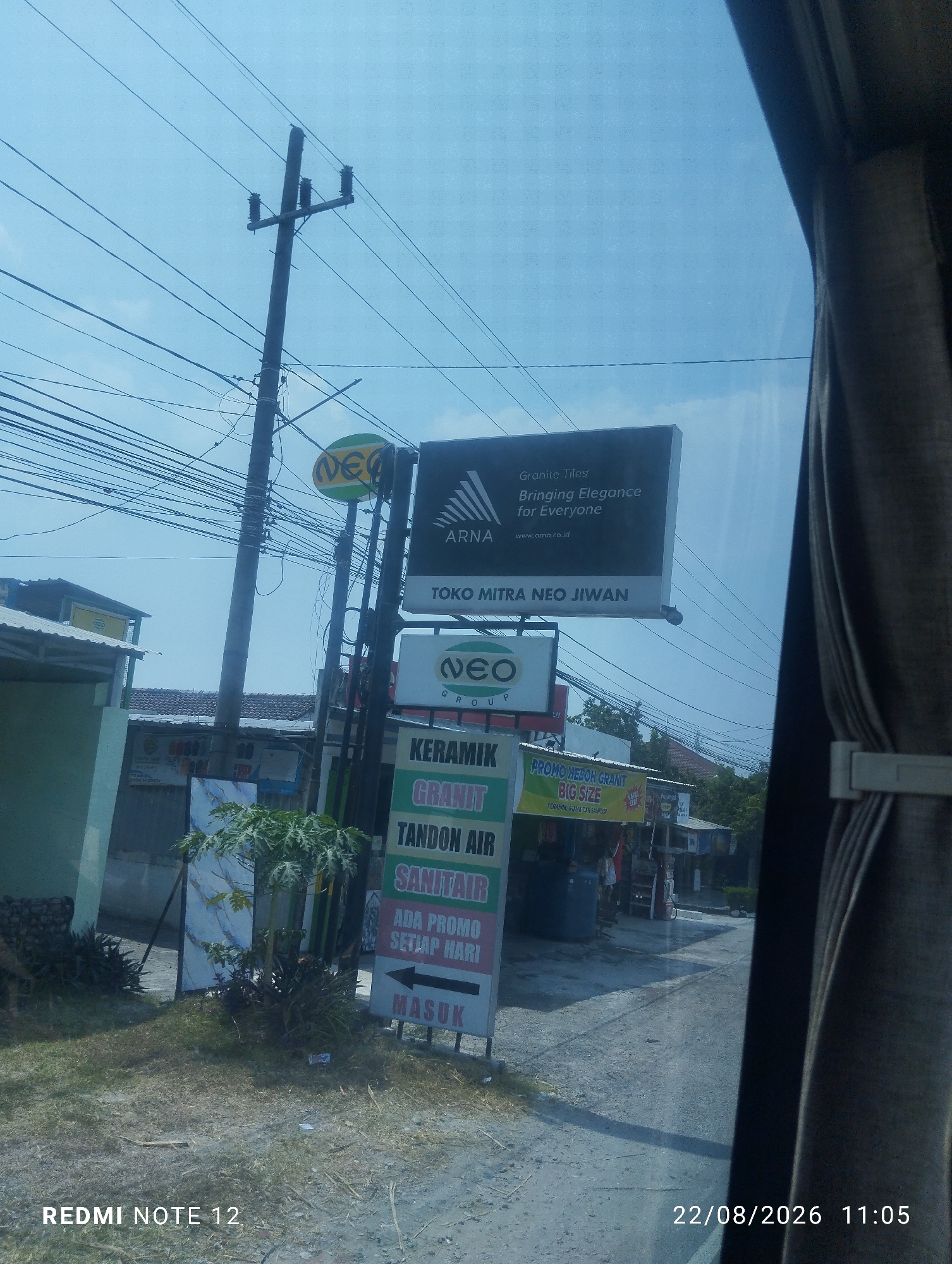The image captures a street scene in Jiwan, Indonesia, under a clear sky. A prominent utility pole with numerous wires crisscrosses the frame, dominating the upper left quadrant. To the right, a large billboard displays "ARNA Granite Tiles, Bringing Elegance for Everyone" with a website address. Below this, another sign for "NEO GROUP" is visible. Further down, a vertical sign lists "KERAMIK GRANIT TANDON AIR SANITAIR" and "ADA PROMO SETIAP HARI," with an arrow pointing to the left indicating "MASUK" (enter). To the left of the signs, a small building with a white roof and green walls is partially visible. In the foreground, a patch of dry grass and dirt covers the ground. A small tree with green leaves stands between the signs and the building. The bottom of the image shows a timestamp reading "22/08/2026 11:05" and "REDMI NOTE 12." The overall scene suggests a commercial area with various businesses advertised along the roadside.