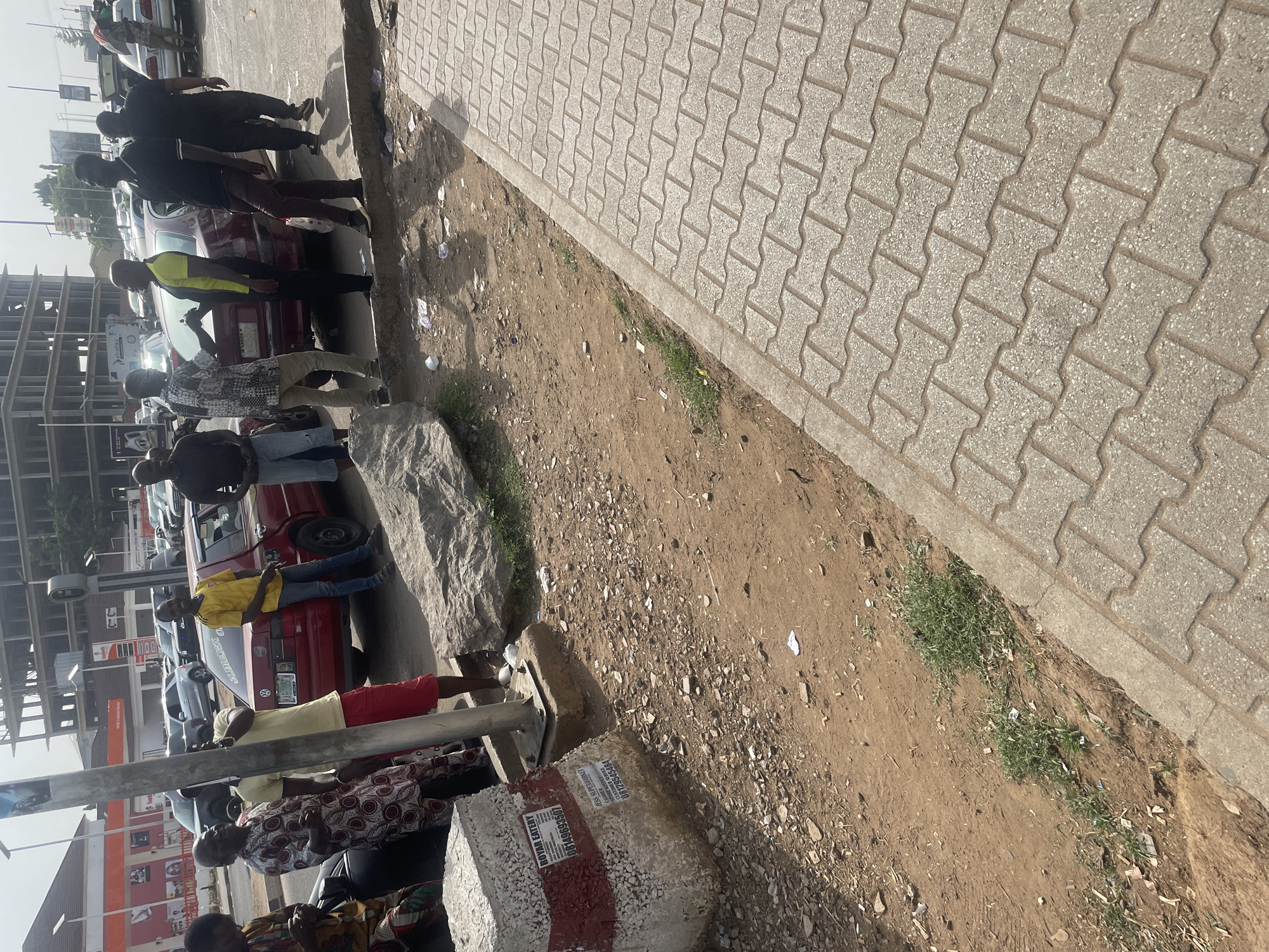 This image captures a vibrant street scene in Abuja, Nigeria, under bright, clear daylight. The perspective is angled downwards, showing a paved sidewalk on the right, a dirt patch in the middle, and a street with parked cars and people on the left.
The sidewalk is constructed from light-colored, interlocking concrete pavers, running diagonally across the upper right portion of the frame. Adjacent to the sidewalk is a strip of reddish-brown dirt, sparsely covered with green weeds and scattered with litter, including small pieces of paper and plastic.
On the left side of the frame, several people are gathered around a few parked cars. The most prominent vehicle is a red sedan, possibly a Volkswagen, with a visible license plate that is partially obscured but appears to be Nigerian. Another silver car is parked behind it. A group of individuals, predominantly men, are standing near the cars, appearing to be engaged in conversation or waiting. One man in a yellow and black safety vest stands near the red car, possibly holding a phone. Other people are dressed in casual attire, including patterned shirts and t-shirts. In the foreground, closer to the viewer, are more individuals, with two women dressed in traditional patterned fabrics visible towards the bottom.
In the bottom left, a large, light-colored concrete block is visible, and another smaller concrete block or barrier features visible text. One sticker reads "ROYAN BATTERY" with a phone number "08149995577". Another sticker on the same block advertises "ESS SUPERMARKET" with the number "08129995586", and a third partially visible number "08123000557".
In the background, behind the parked cars and people, an unfinished multi-story building with exposed concrete and scaffolding suggests ongoing construction. Further in the distance, what appears to be a gas station or commercial establishment sign is partially visible, showing "SIGNAL" and hints of other branding. The overall atmosphere suggests a typical busy urban environment with daily activities taking place.