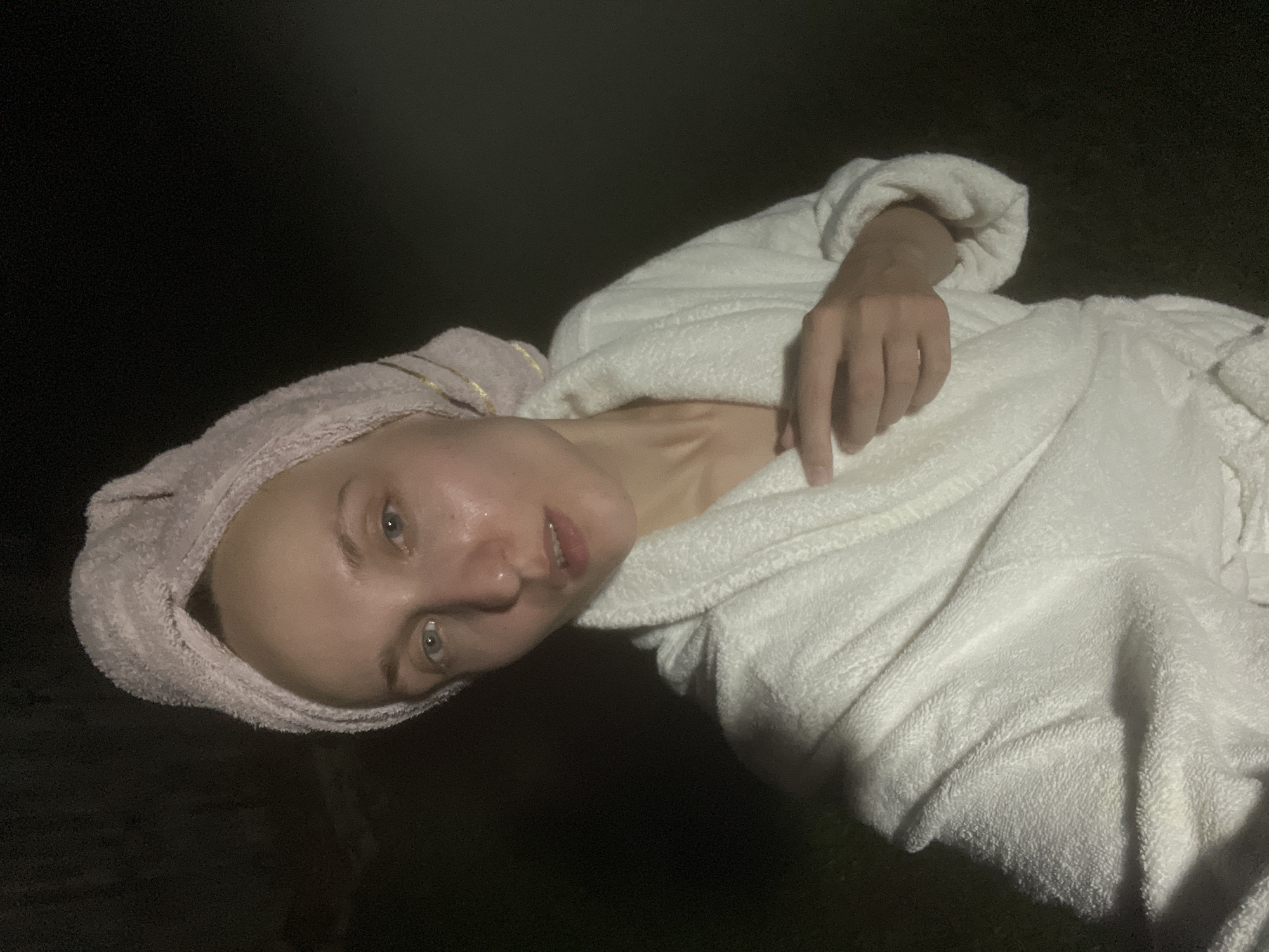 A woman with a pink towel wrapped around her head and a white bathrobe is standing outdoors in the dark. Her face is partially illuminated, showing her eyes, nose, and mouth. Her skin appears to be slightly moist. She is holding her hand to her chest. The background is dark, suggesting it is nighttime. There is no indication of the city of Sharkan, Russia in the image.
