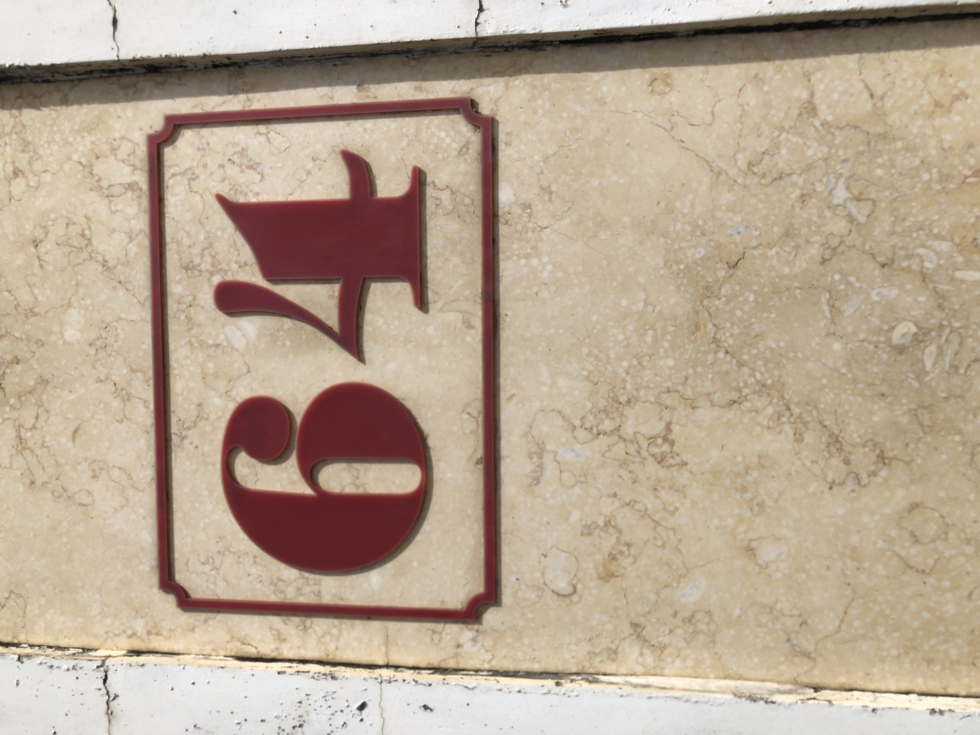 A close-up, eye-level shot shows a house number "64" on a beige, marble-patterned wall. The numbers are a deep red color and are encased in a darker red, ornate rectangular frame. The wall is bordered on the left by a white, textured surface with visible cracks and a darker edge. The right side of the wall is bordered by a dark, rough surface. The lighting suggests it is daytime, casting subtle shadows from the numbers.