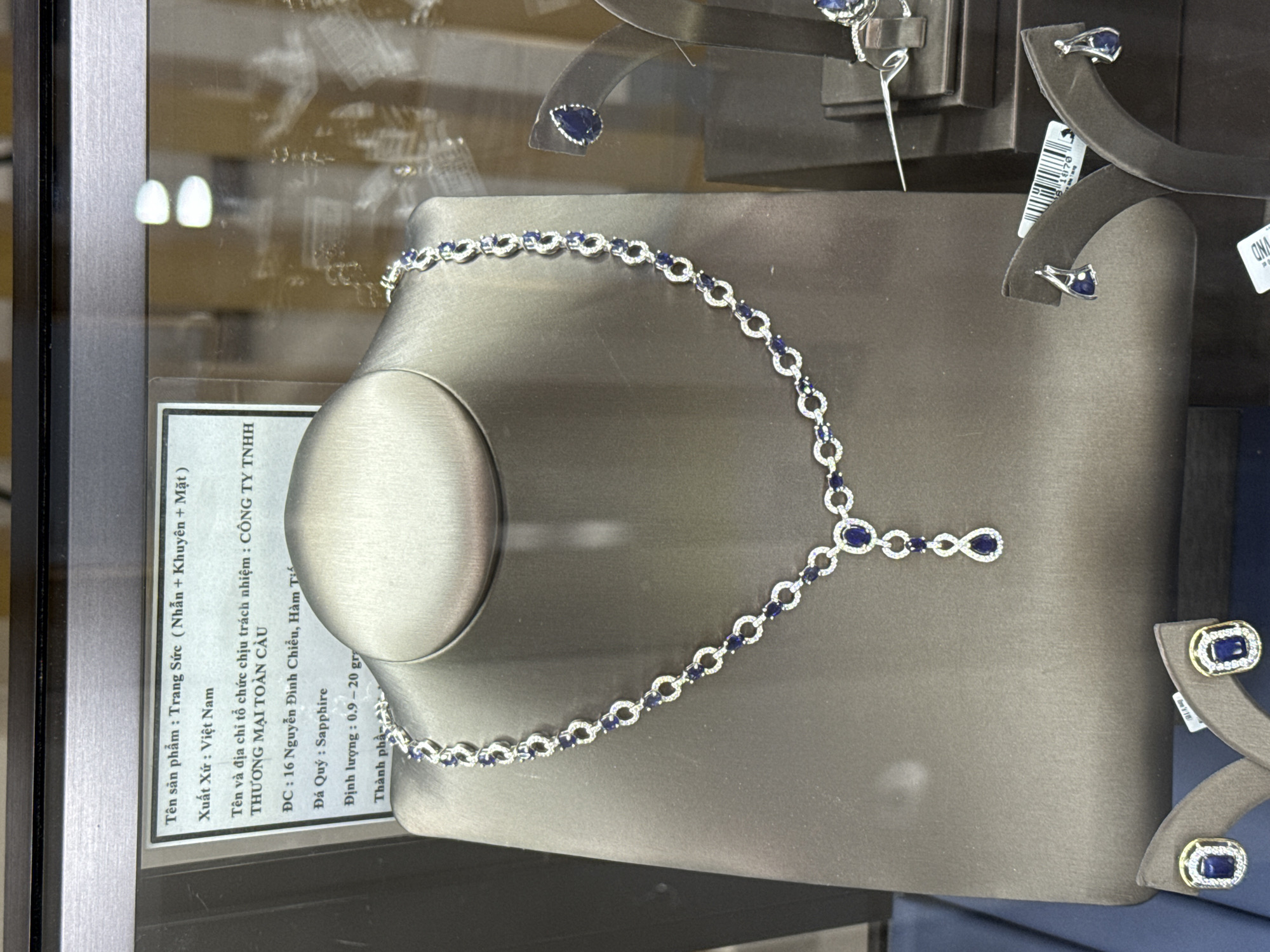 The image captures a jewelry display, likely within a shop in Nha Trang Ward, Vietnam. The display showcases a necklace with silver settings and sapphire stones, arranged on a gray, mannequin-like stand. A pair of earrings with similar sapphire and silver design are displayed on stands behind the necklace. A small sign provides information about the product, including the name "Trang Sức (Nhẫn + Khuyên + Mặt)," its origin ("Việt Nam"), and the name of the responsible organization: "CÔNG TY TNHH THƯƠNG MẠI TOÀN CẦU." The sign also mentions the address, "16 Nguyễn Đình Chiểu, Hàm T," and specifies the gemstone as "Sapphire" with a weight of "0.9-20 gr."