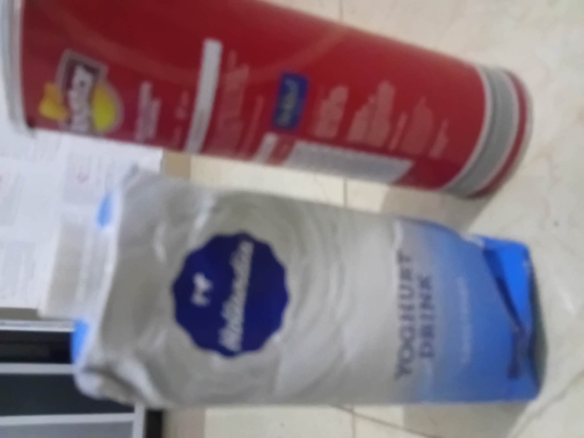 The image shows a red can of Pringles and a white and blue carton of Muller Yoghurt Drink on a tiled surface. The location is likely Akwete, Nigeria.
