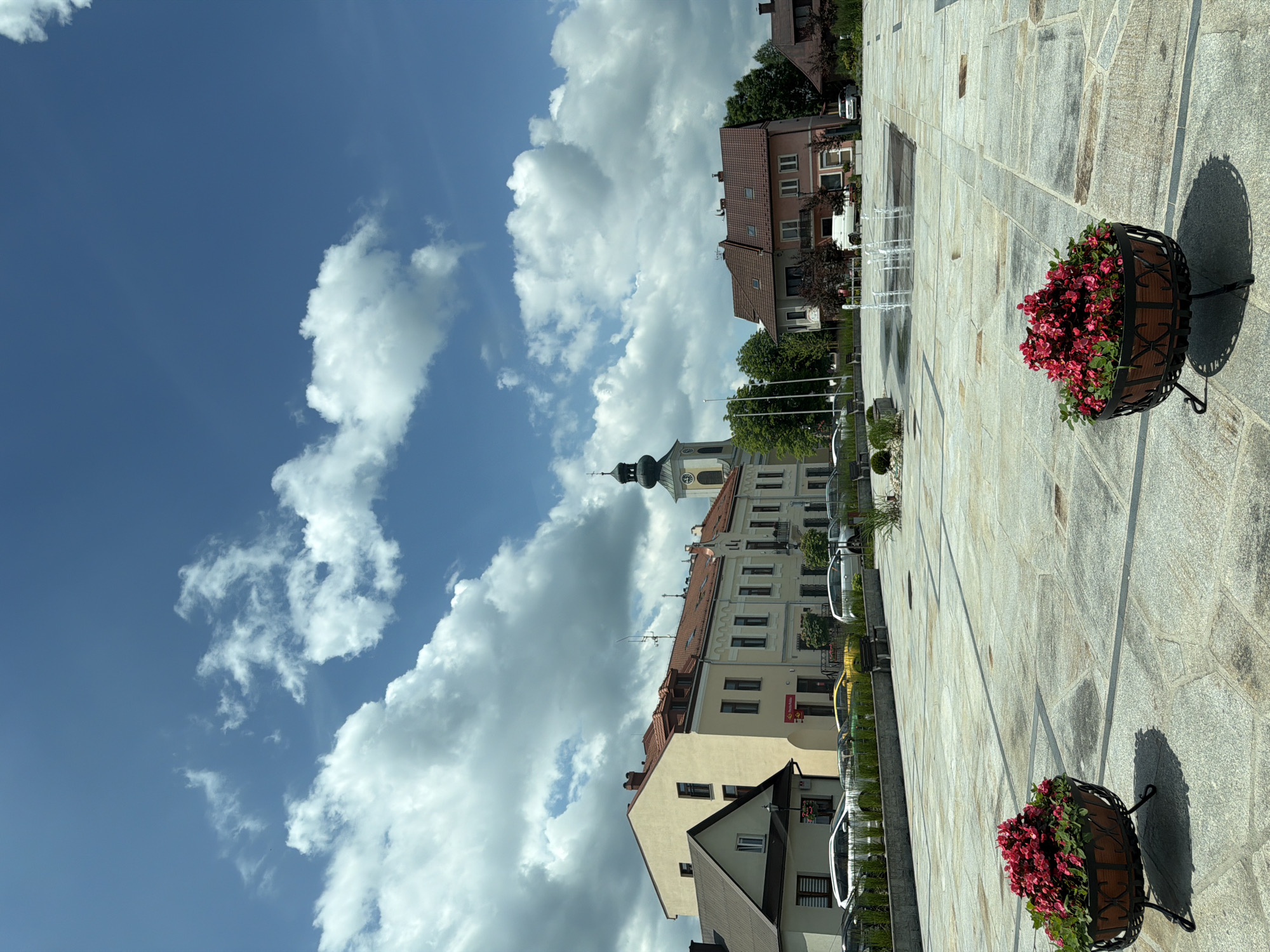 The image shows a sunlit town square in Tymbark, Poland, on a partly cloudy day. In the foreground, two large, dark planters filled with bright red flowers on black metal stands are placed on a paving stone surface. The paving stones are large and rectangular, laid in a pattern. Further into the square, a modern fountain with several water jets is active. In the middle ground, a row of buildings lines the square, featuring varying architectural styles and colors, including a pale yellow building with a red-tiled roof and a building with a dark green spire and a clock face. Trees and bushes are visible between the buildings and in planters. Several cars, including a white van and two silver cars, are parked along the street behind the buildings. In the background, more buildings and trees are visible under a bright blue sky with scattered white clouds. The overall scene is a peaceful, well-maintained urban environment.