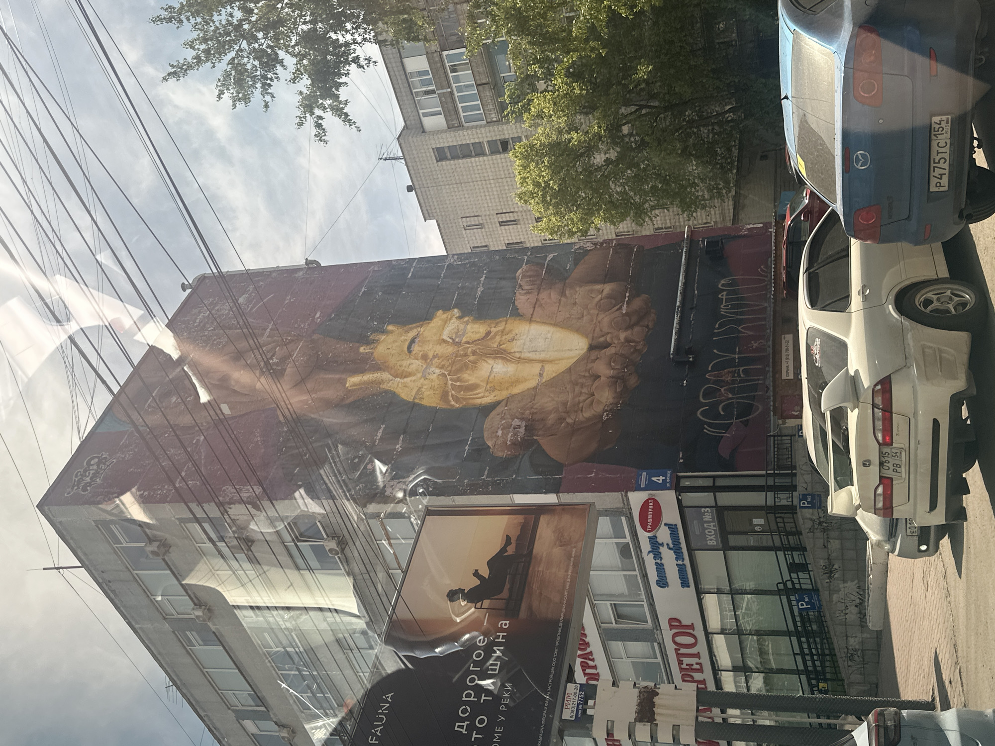 A street scene, likely in a Russian city, is captured during bright daylight from inside a vehicle, evidenced by reflections across the image. The central feature is a large, multi-story building adorned with a prominent, stylized mural. The mural depicts a golden-hued, possibly disembodied head or bust, with hands, set against a dark background, evoking an artistic or classical style.
Below the mural, various storefronts and signs are visible on the building. A large billboard shows a silhouette of a person sitting on a chair with text "дорогое то тишина" (expensive to silence/quiet). Other signs include "ТРАВМПУНКТ" (Traumatology clinic/First aid post), "ВХОД №3" (Entrance No. 3), and "PETOP". Graffiti reading "GRATE WIND" is also visible on the lower part of the mural wall.
Two cars are parked on the street to the right: a blue Mazda with license plate "P475TC 154 RUS" (indicating Novosibirsk region, Russia) and a white car with a prominent spoiler and license plate "0615 PB 54 TN" (with 54 also being a Novosibirsk region code). Numerous utility wires crisscross the bright, cloudy sky above the buildings. Another multi-story building is partially visible behind the main structure.
Visible text includes: "FAUNA", "дорогое то тишина", "ОМЕ У РЕКИ", "RA&FAUNA (ФЛОРА И ФАУНА) ЗАСТРОЙЩИК ООО "СИК" ПРОЕКТНЫ", "РИМ", "8(383)217-00-20", "№ 7752", "ΟΓΡΑΦ", "PETOP", "ТРАВМПУНКТ", "Ваше здоро пиша забота!", "ВХОД №3", "4", "P", "GRATE WIND", "ОХРАНА: +7 (913) 700-51-32", and the license plate numbers "P475TC 154 RUS" and "0615 PB 54 TN". The phone numbers and license plates indicate a Russian location, specifically the Novosibirsk area.