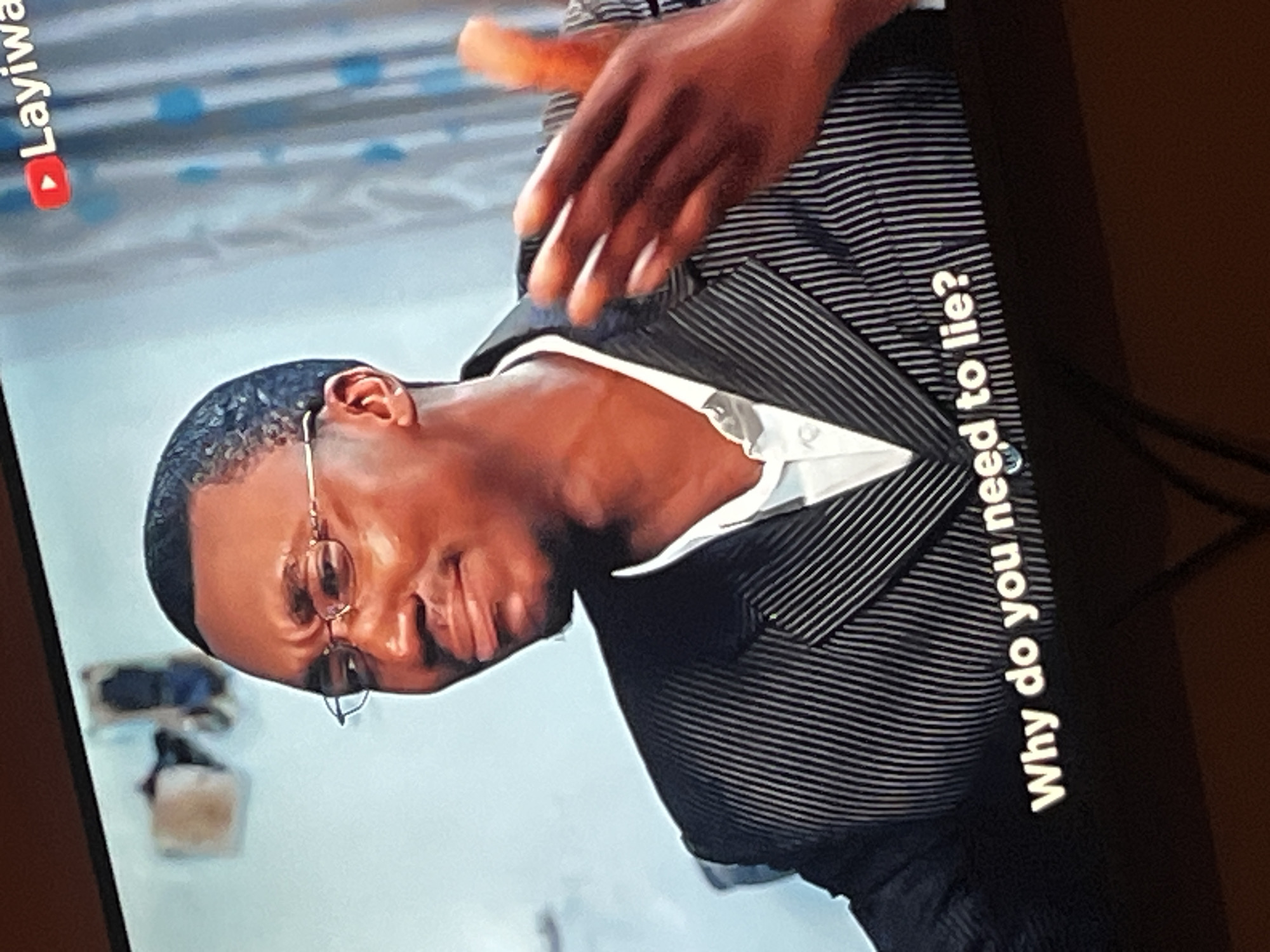 A man in glasses and a pinstripe suit, with a white shirt collar, is speaking with his right hand raised. A YouTube logo and text that reads "Layiwa" are visible in the upper right corner of the image. Below the man, a subtitle reads "Why do you need to lie?". The background consists of a light blue wall with a vertical striped curtain. In the upper left corner, a light-colored object with a dark base is mounted on the wall. The image appears to be a still from a video being viewed on a screen.