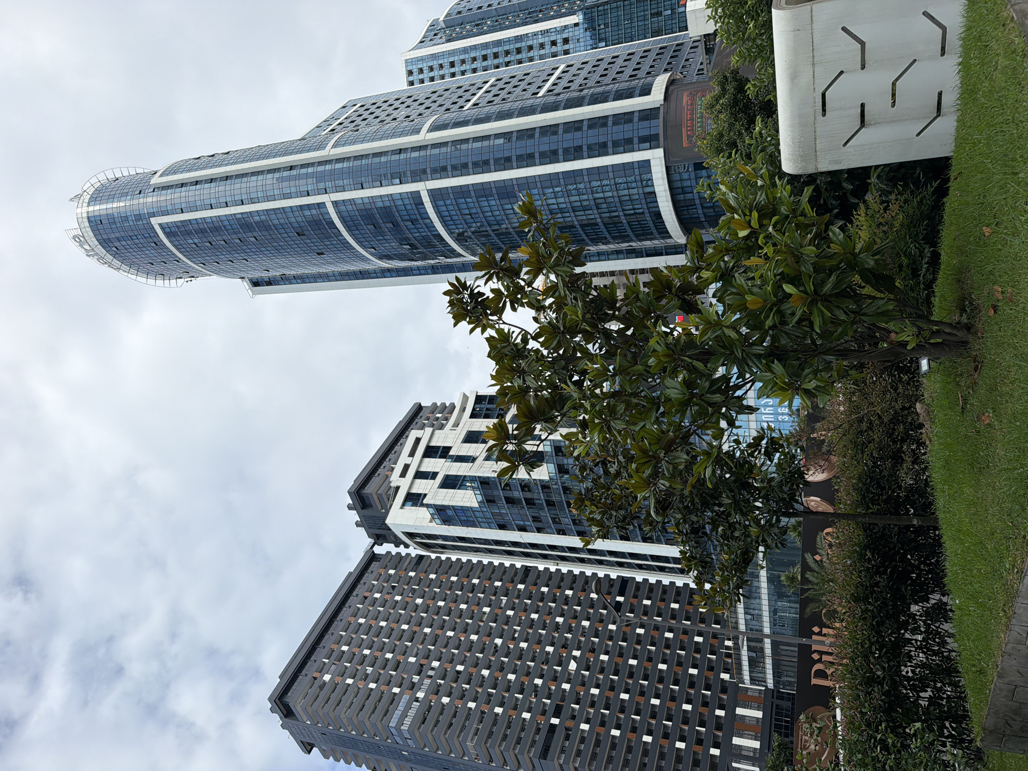 The image shows a view of modern high-rise buildings in Batumi, Georgia. In the foreground, there are green trees and manicured bushes with a grassy area and a concrete structure. Behind the greenery, several tall buildings are visible. The tallest and most prominent building on the right is a curved skyscraper with a glass facade and white vertical accents. To its left, there is another tall building with a white and blue patterned facade, and further to the left, a large dark-colored building with numerous balconies and orange accents. The sky is overcast. The overall scene depicts an urban environment with contemporary architecture.