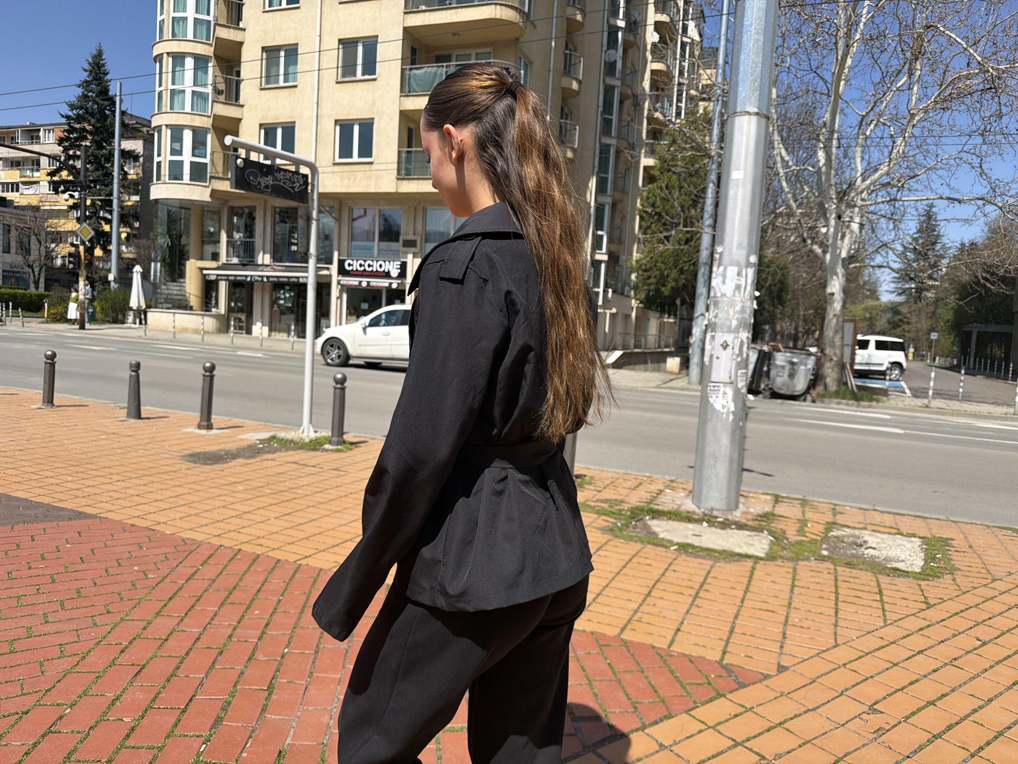 The image depicts a young woman with long brown hair styled in a ponytail, seen from the back, walking on a brick sidewalk. She is wearing a black suit with a belted jacket. The setting appears to be an urban street in Sofia, Bulgaria, evident from the architectural style of the buildings, the presence of street signage with Bulgarian text, and the 'Ciccone' storefront. A white car is parked across the street. A streetlight stands on the sidewalk, and the street is visible with occasional vehicles.