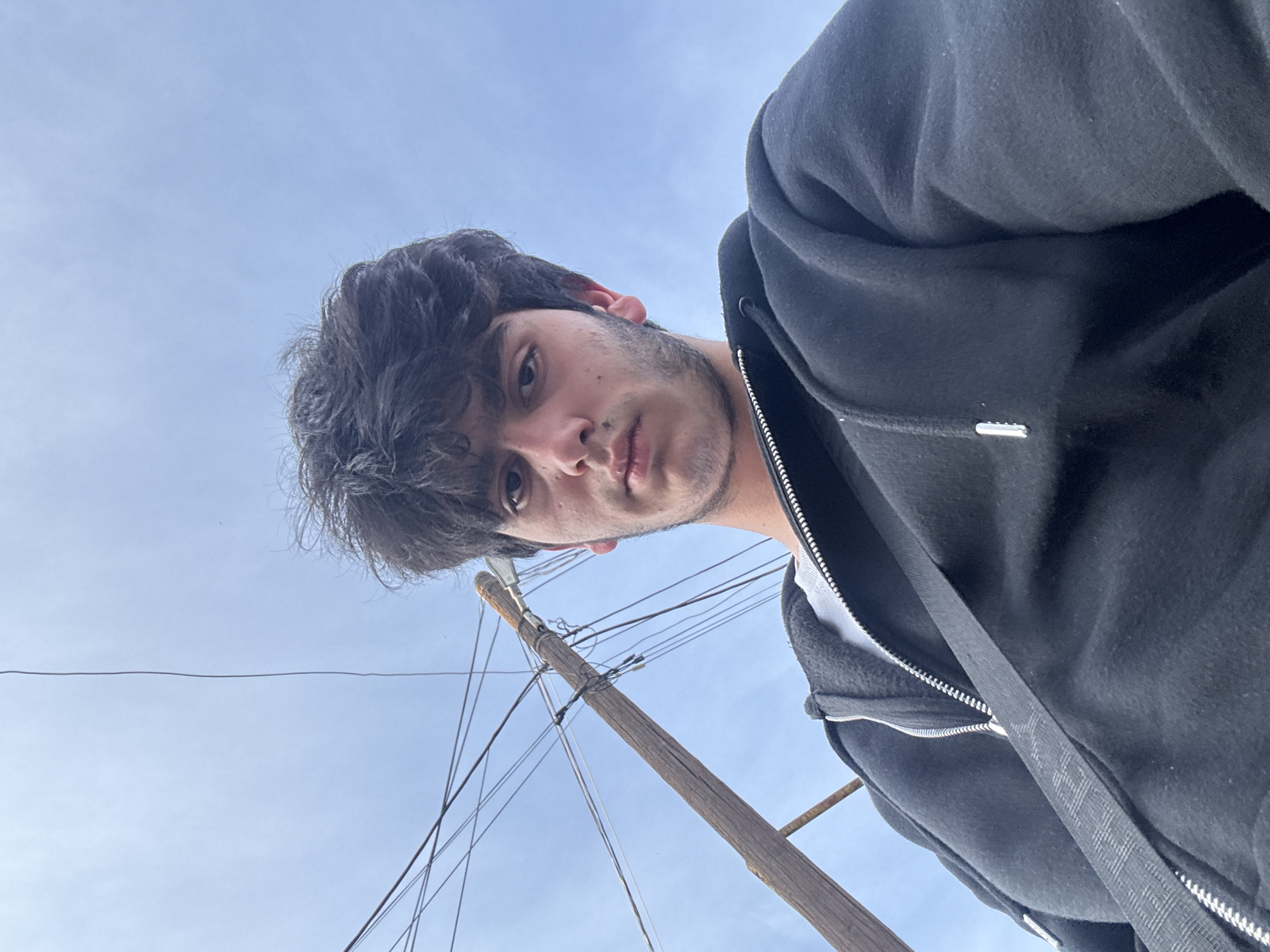 A low-angle selfie photograph features a young man with dark, curly hair, light skin, and stubble. He is wearing a black zippered hoodie over a white shirt. Behind him is a clear, light blue sky. A wooden utility pole with several wires running from it is partially visible in the background.
Due to the limited information available in the image, it is not possible to determine the exact location within Yerevan, Armenia.